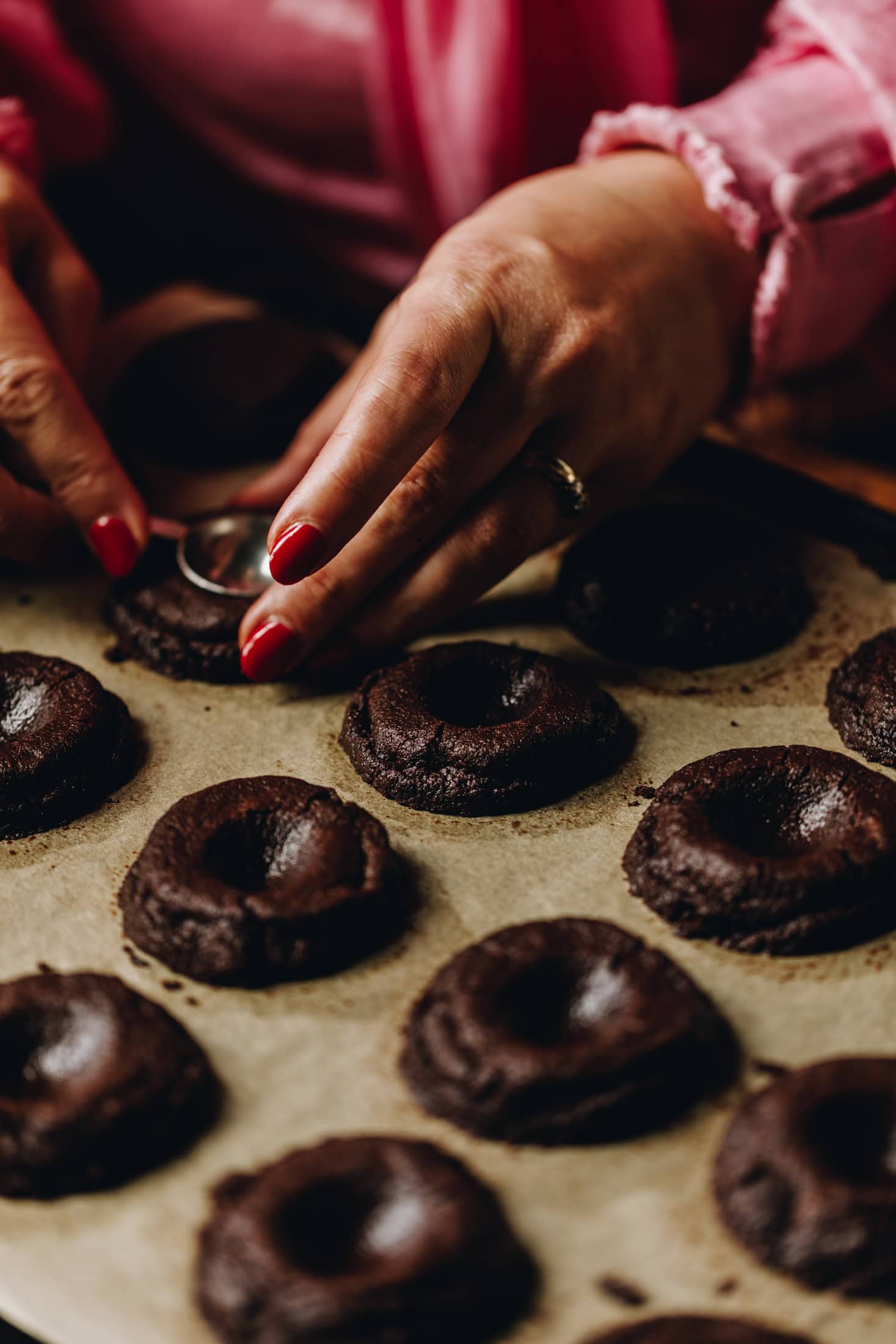 On a tray lined with baking paper is freshly baked Buckeye cookies. They have been indented with a teaspoon and one cookie is being indented with a teaspoon in the photo.