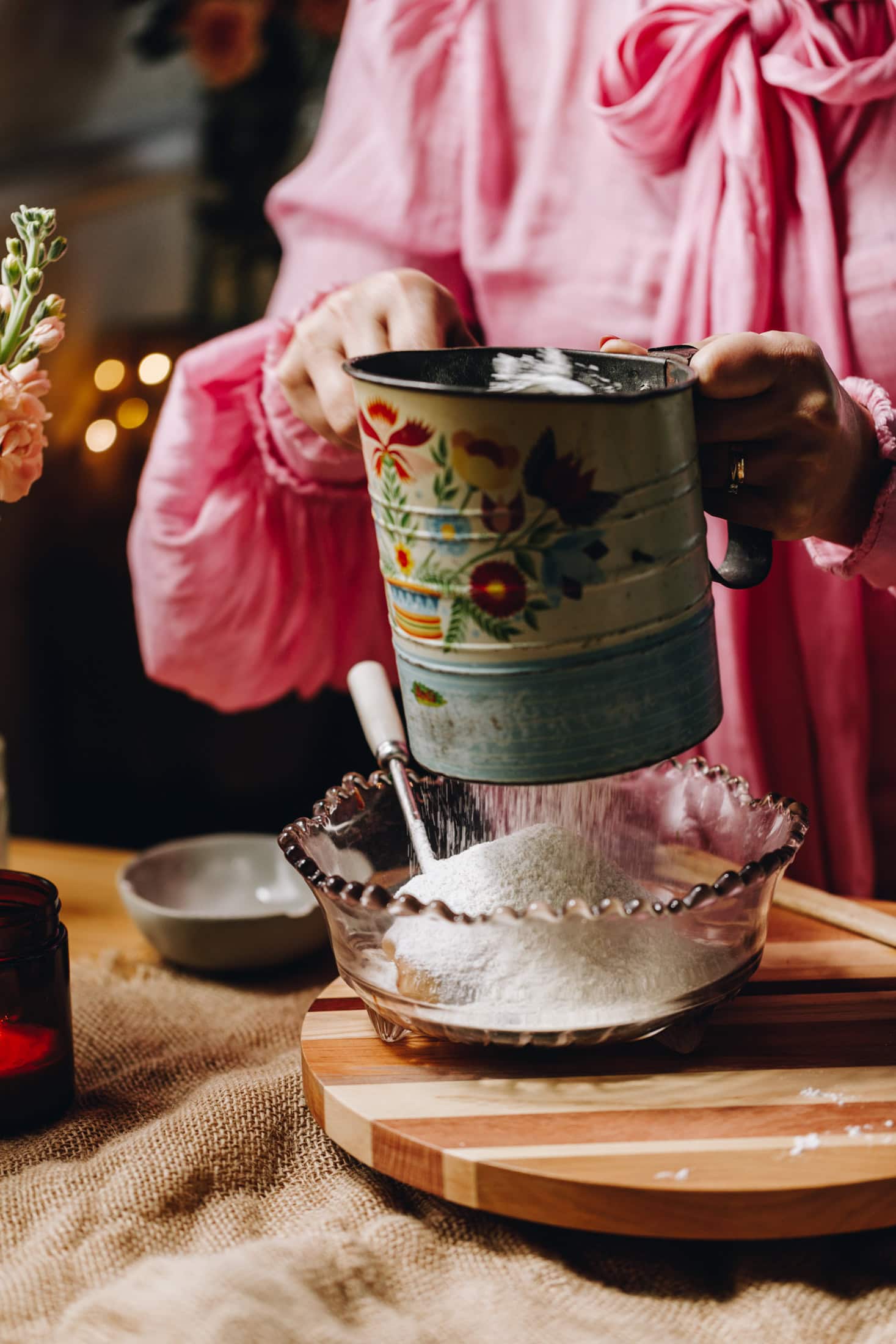 On a wooden board sits a purple vintage bowl. In it is sifted dry ingredients that Naomi is sifting in to the bowl with a blue vintage sifter.