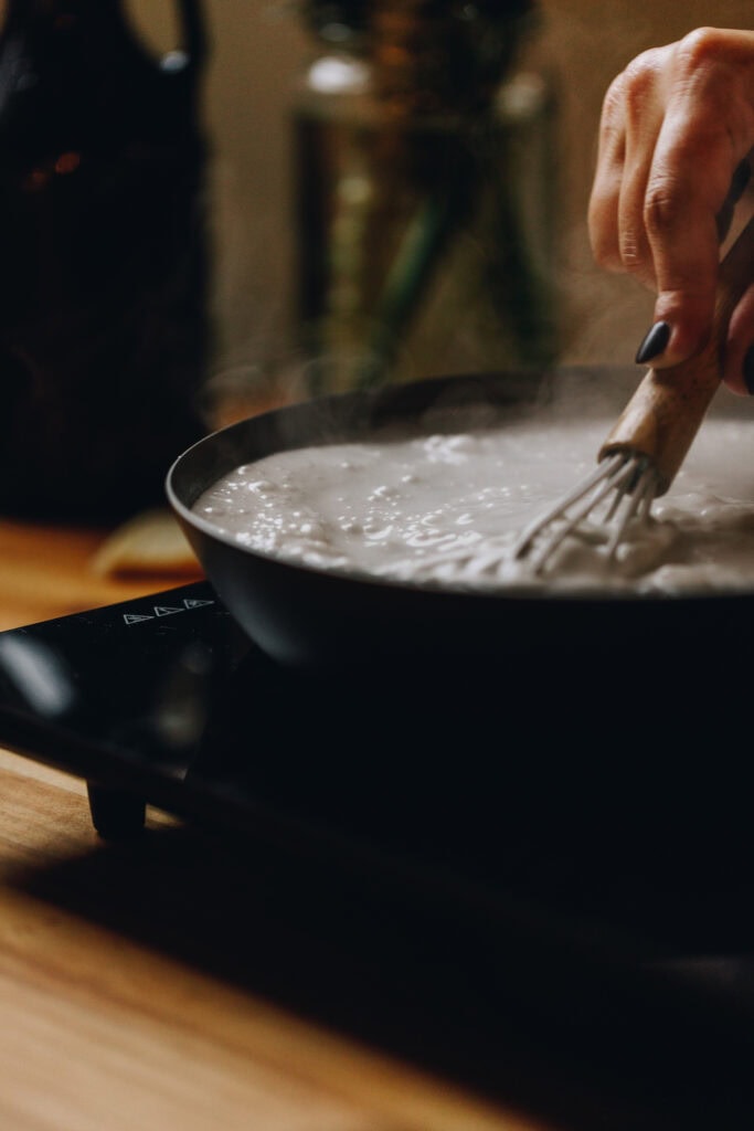 Close up of the Coconut Buns (Pani Pop) sauce boiling in a frying with a small whisk stirring the mixture. It sits on a portable stove top on a wooden table. 
