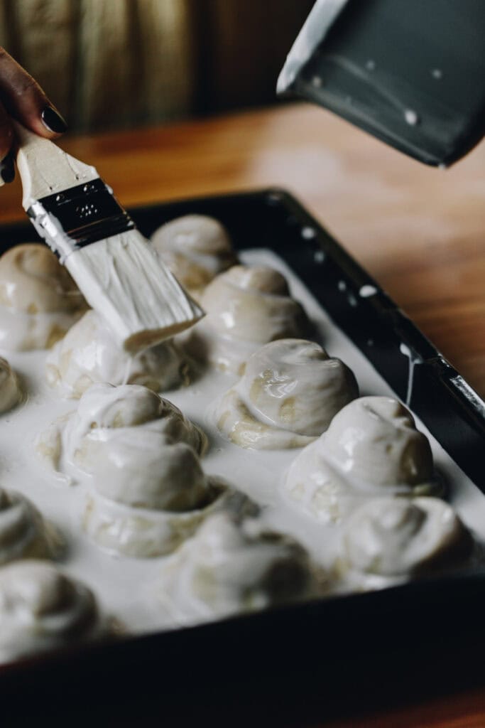 Close up of the unbaked Coconut Buns (Pani Popo) being bathed in coconut sauce before baking, they are in a black metal tray on a wooden table. 