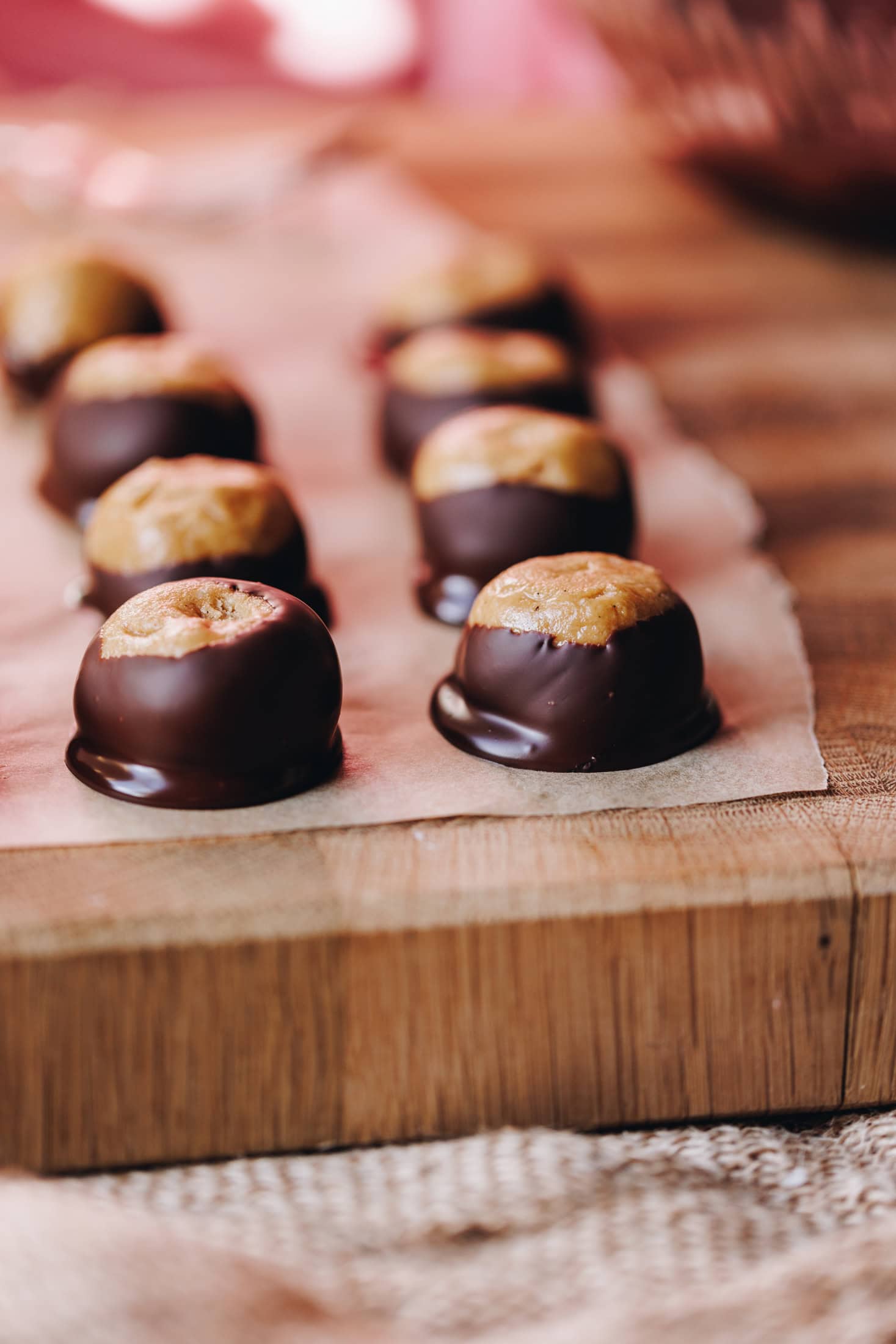 A wooden board lined with brown baking paper sits on a cloth. On the baking paper is freshly decorated peanut butter buckeye truffles. They truffles are round and they have been dipped in chocolate with the top part of the truffle exposed.