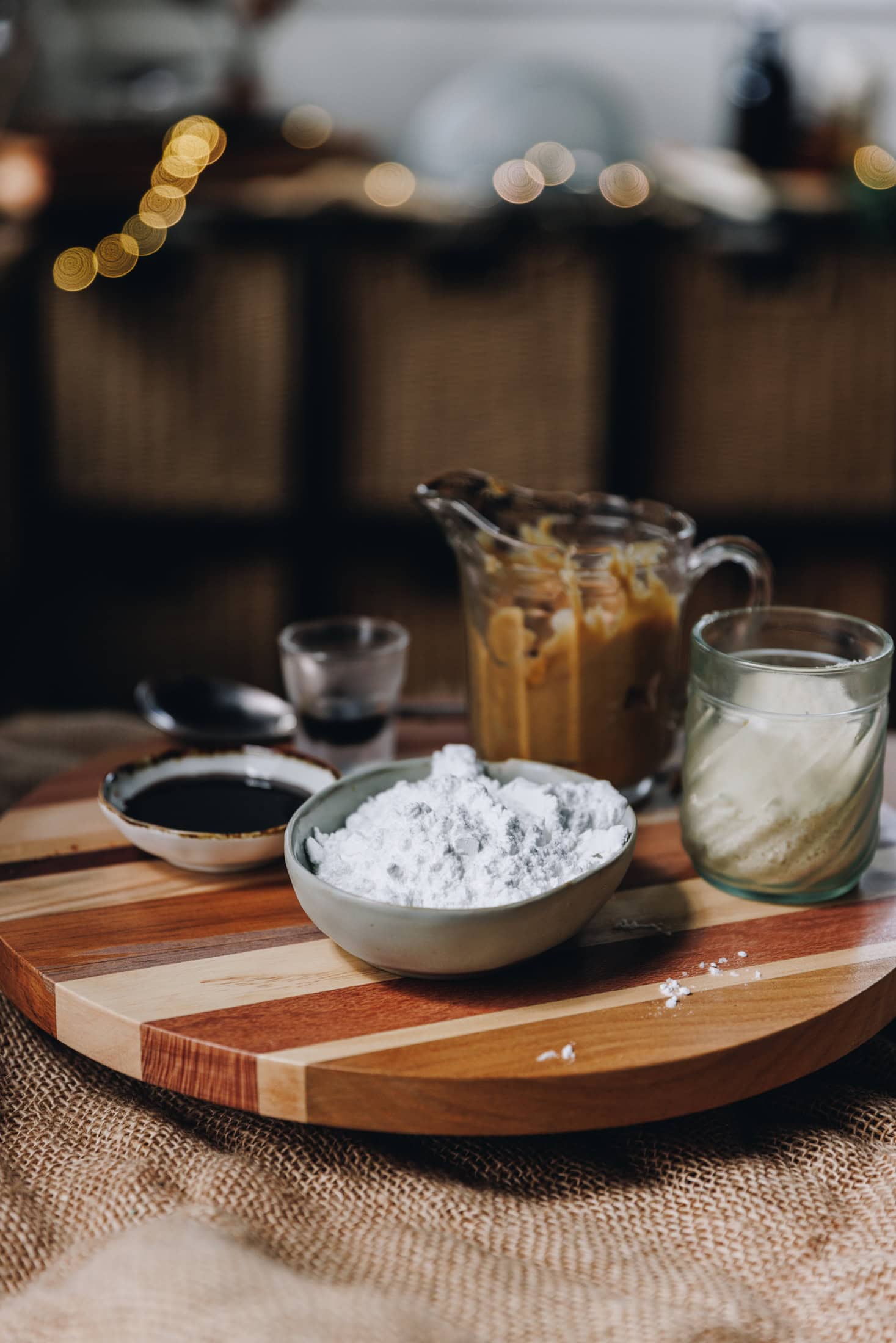 On a wooden board sits different vintage bowls and glasses holding the ingredients for the truffles. There is icing sugar, peanut butter, vanilla paste and almond meal in view. Blurred fairy lights are in the background.