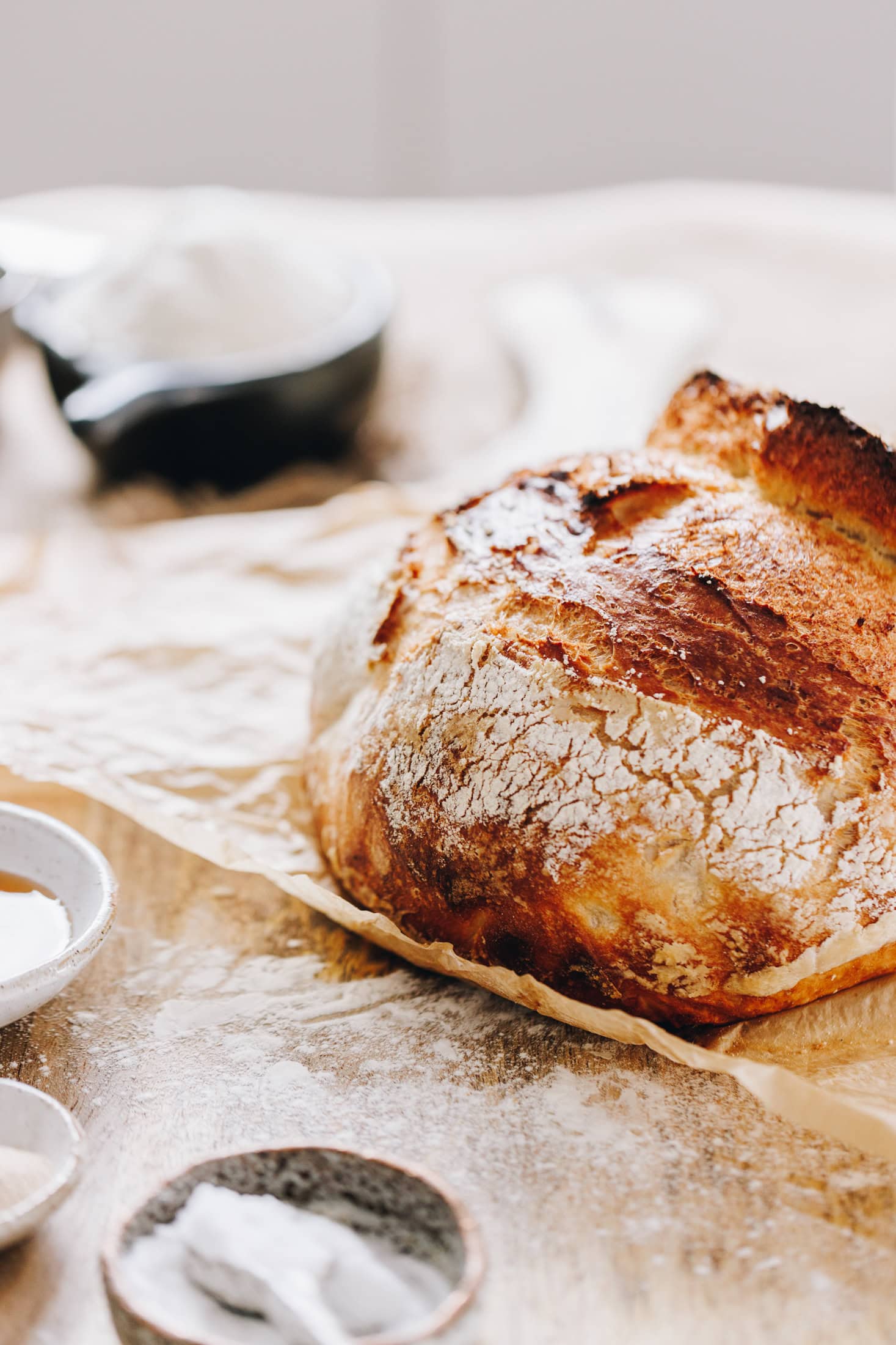 A freshly baked rustic no-knead bread loaf siting on baking paper, on top of tablecloth. It is uncut and shows the crispy bread.