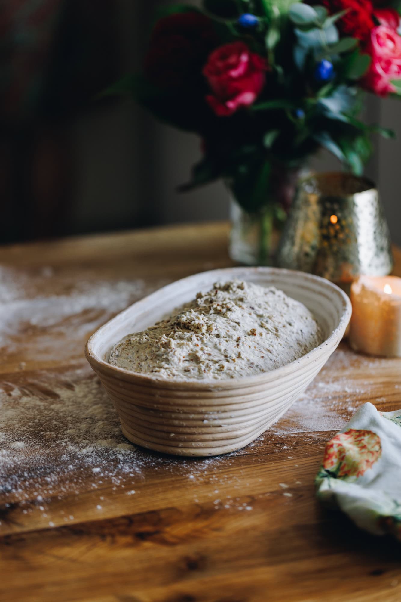 A dough is in a sourdough banetton, it sits on a wooden, floured table with flowers in the background. 