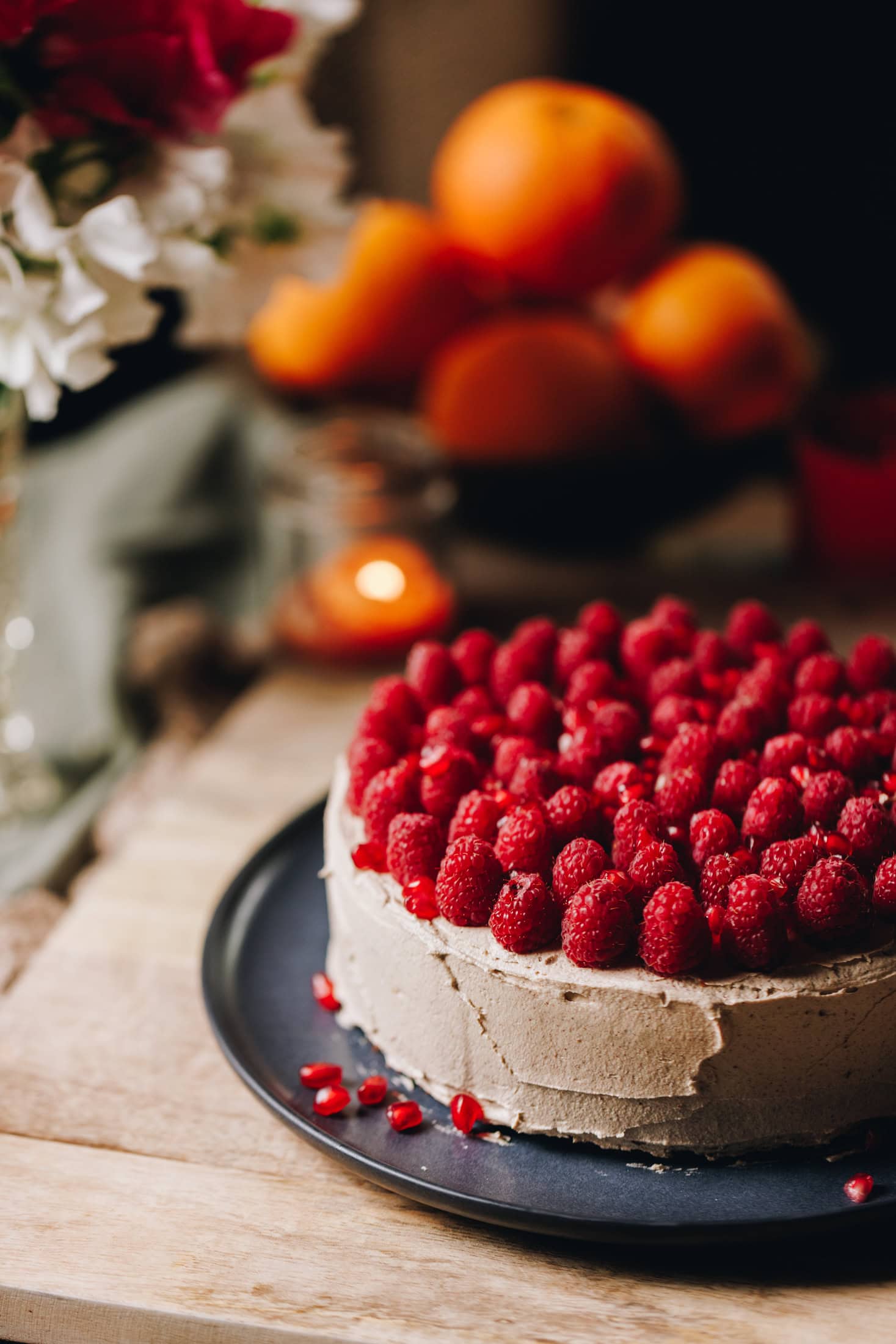 A blue plate sits on a wooden table. On the plate is a whole orange cake that is decorated in simple cinnamon buttercream with fresh raspberries covering the top of the cake with grated orange zest. A bowl of oranges are seen in the background. 
