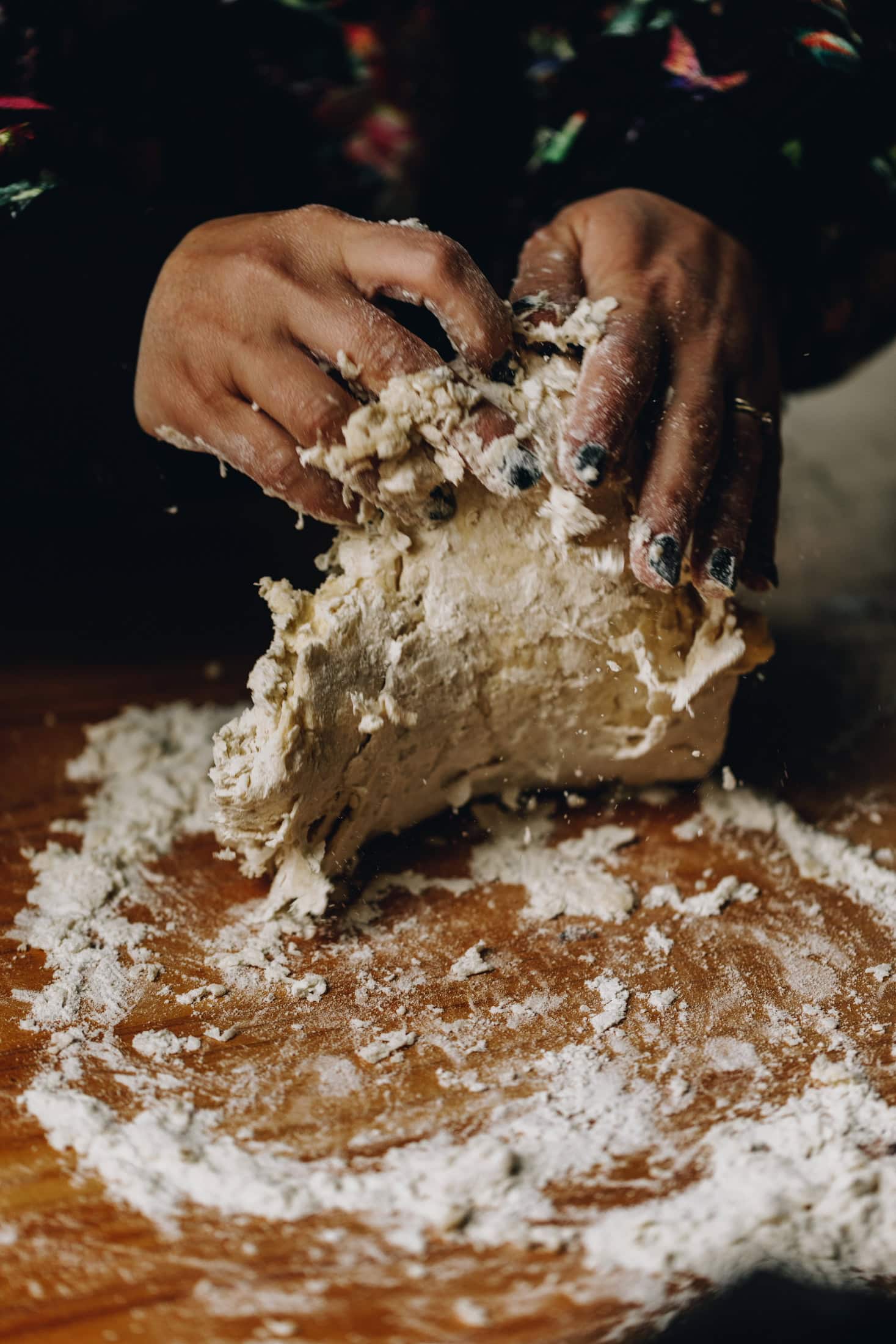 On a wooden floured table, two hands are kneading a small bread dough. The dough is being lifted by the hands. 
