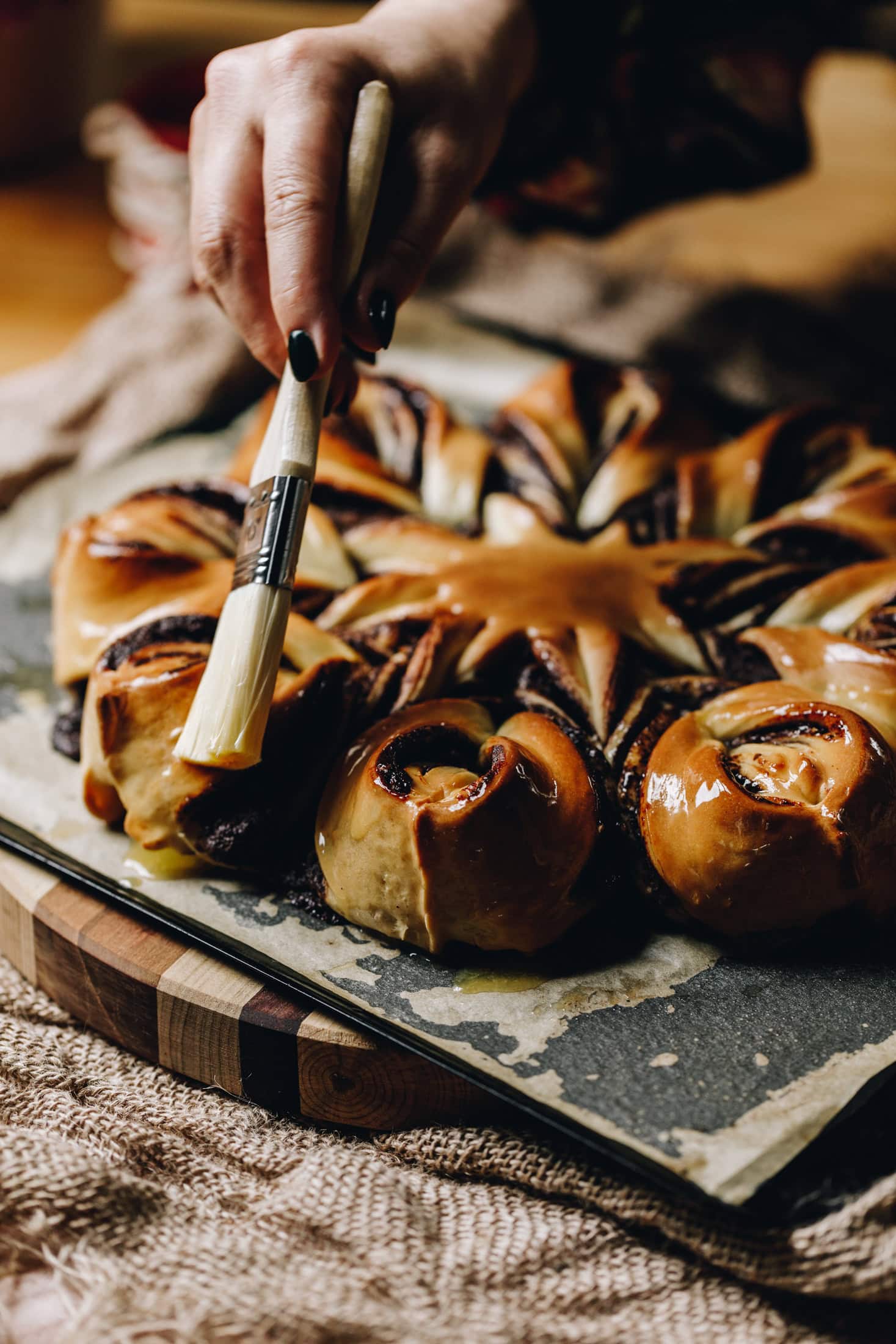 A freshly baked Chocolate Star Bread is on a black tray with brown baking paper. It is being brushed with a glaze with a pastry brush. It is on a wooden table. 