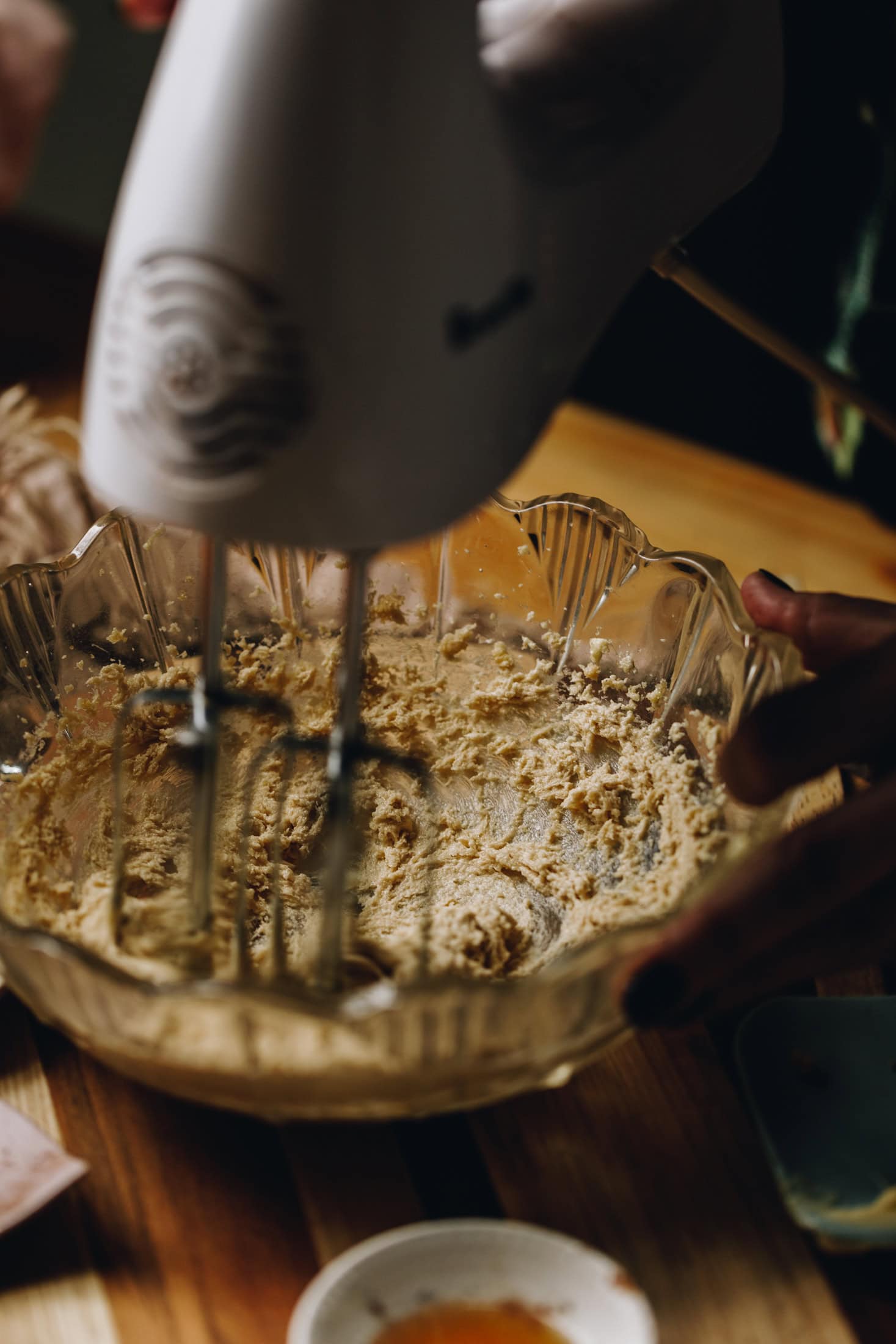 In a vintage glass bowl butter and sugar is being whipped with a white hand mixer. It is on a wooden table. 