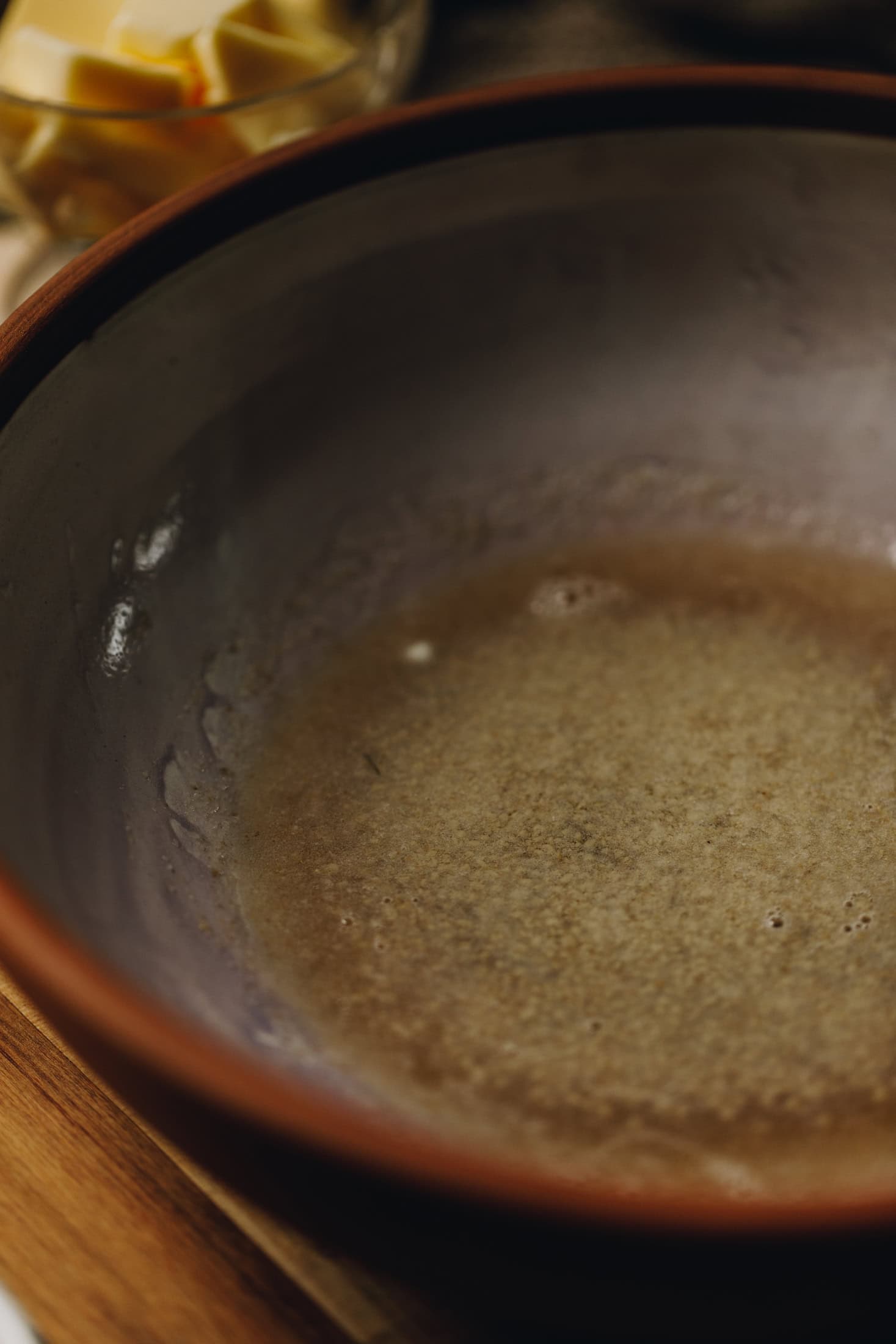 In a small cermic bowl, yeast has activated and it floating to the top of the water. The bowl is on a wooden table. 