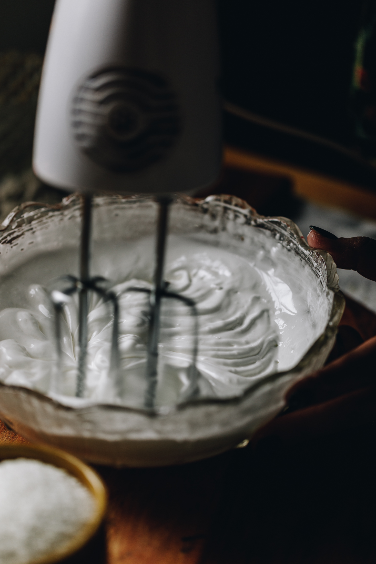 Swiss meringue is being whipped in a glass bowl that is on a wooden table. It is being whipped by a white hand mixer.