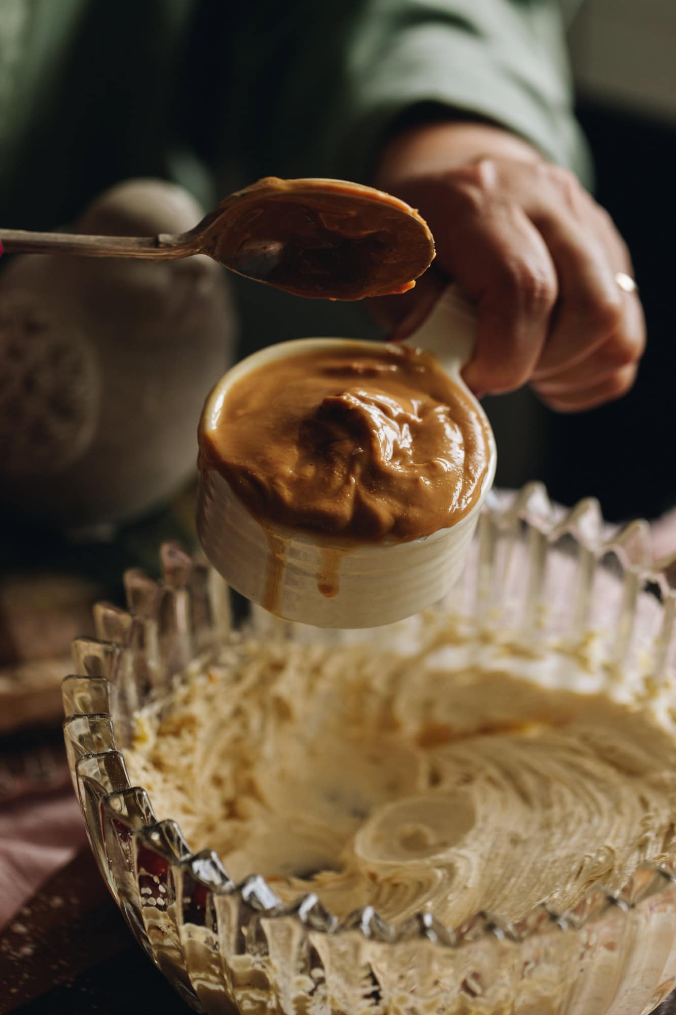 A glass vintage bowl with jagged edges sits on a striped wooden board. In the bowl is whipped butter and sugar. Naomi is holding the peanut butter in a cup above the bowl and has a spoon, ready to tip it in to the bowl.