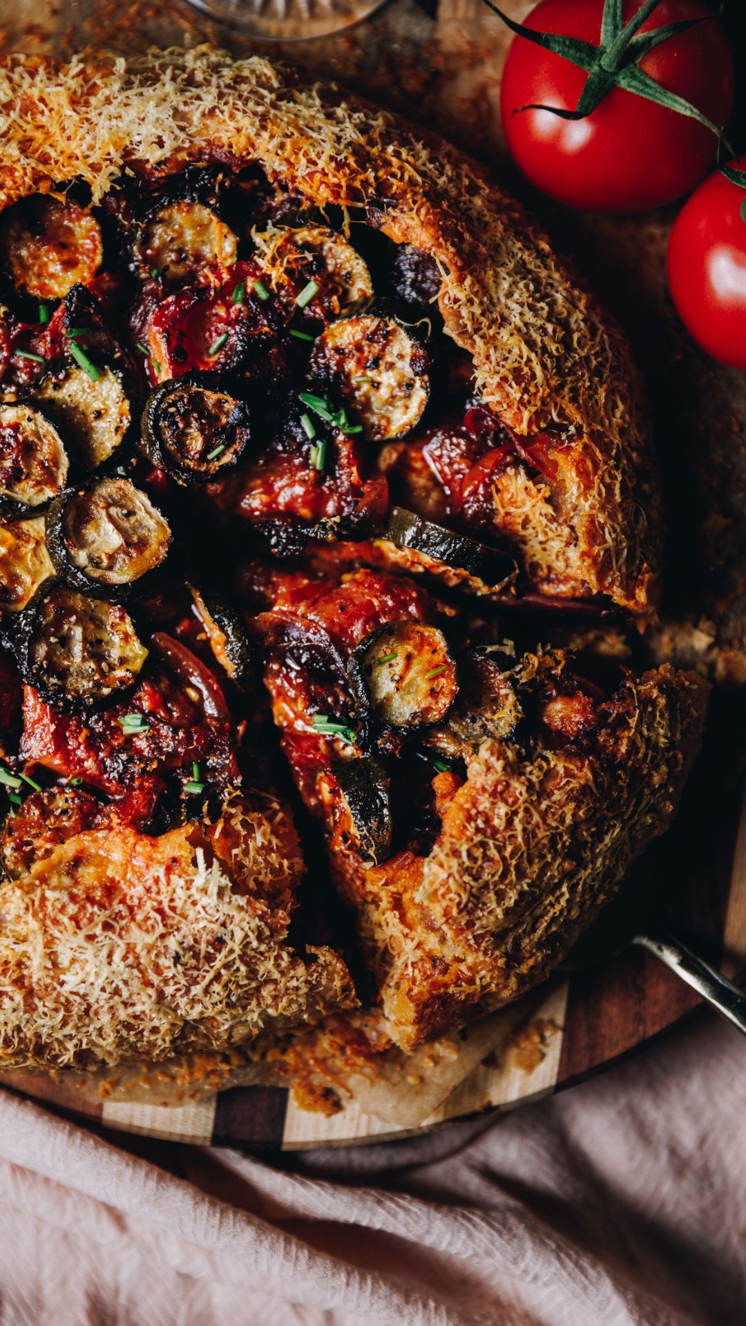 A freshly baked roasted tomato and courgette galette is on a striped wooden, round board. A piece has been cut and the inside of the pie is revealed. There is a pastry layer, a creamy layer and loads of roast vegetables.
