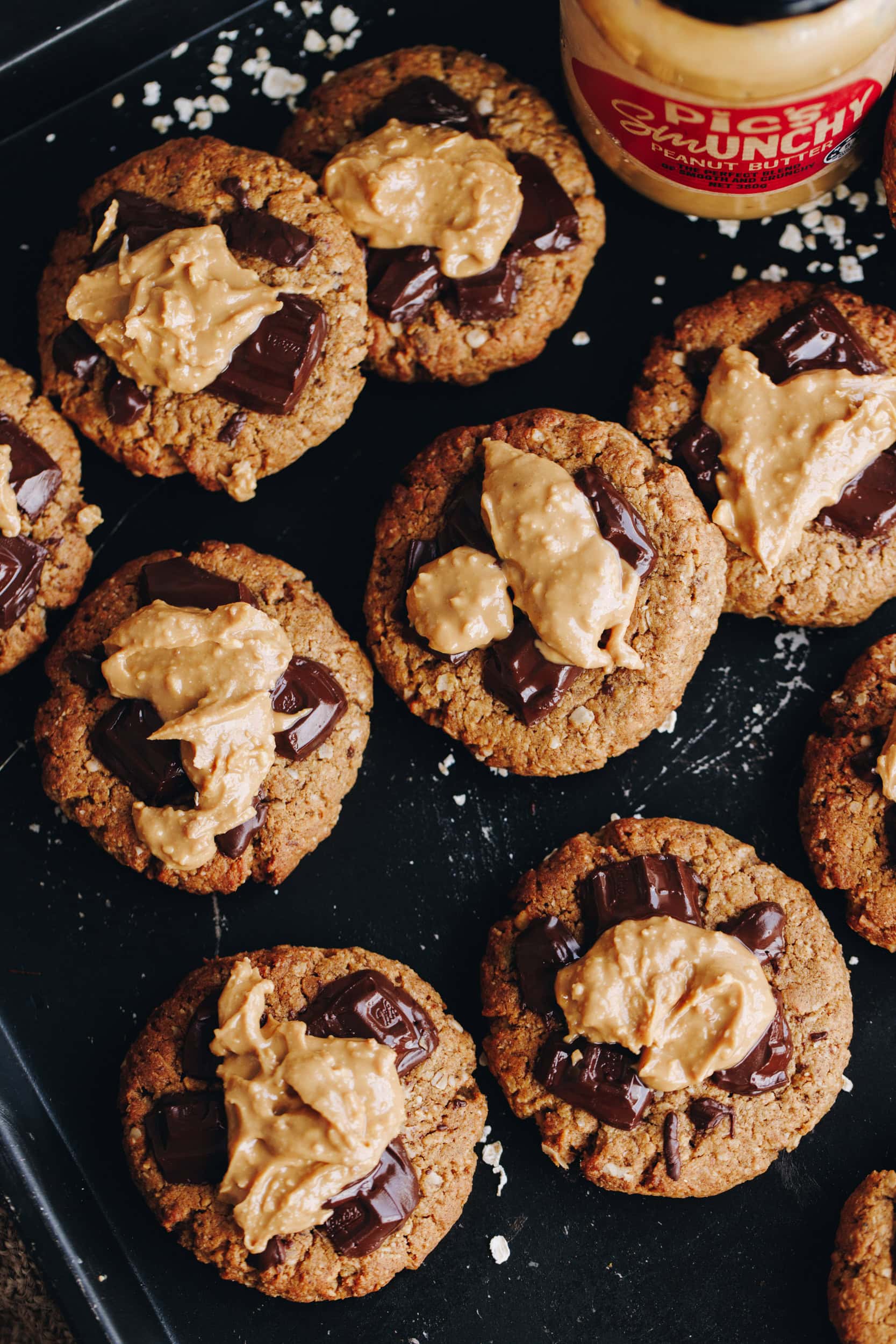 On a black tray are freshly baked wheat-free PB breakfast cookies. Th stop shot shows the cookies with chocolate on the top and peanut butter spread in the centre, there is a scattering of flaky salt on the tray. A Pic's Smunchy peanut butter jar in the corner.