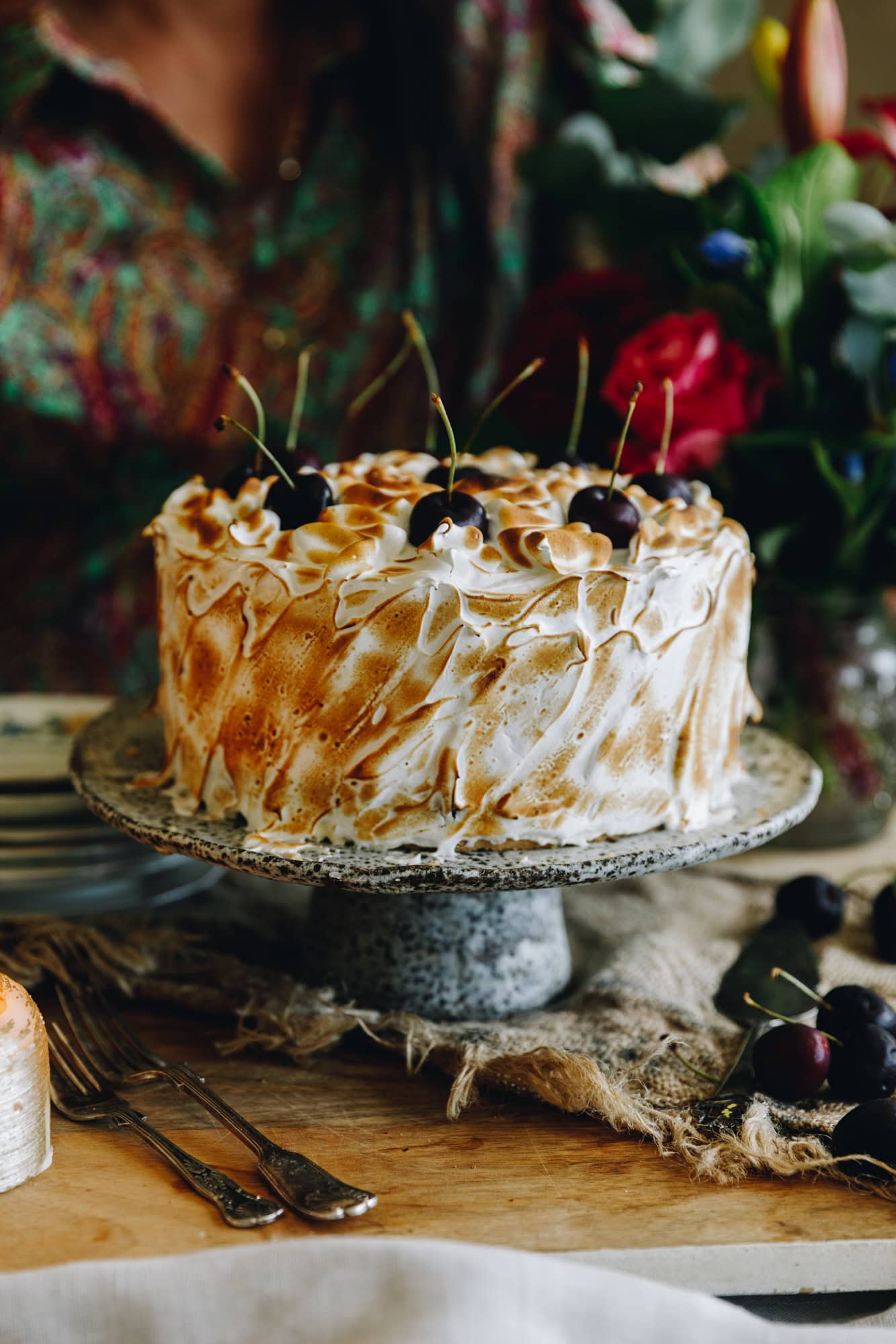 A whole Gluten-free almond sponge is on a ceramic cake stand on a wooden table. It Is covered in torched Swiss meringue and topped with whole cherries. 