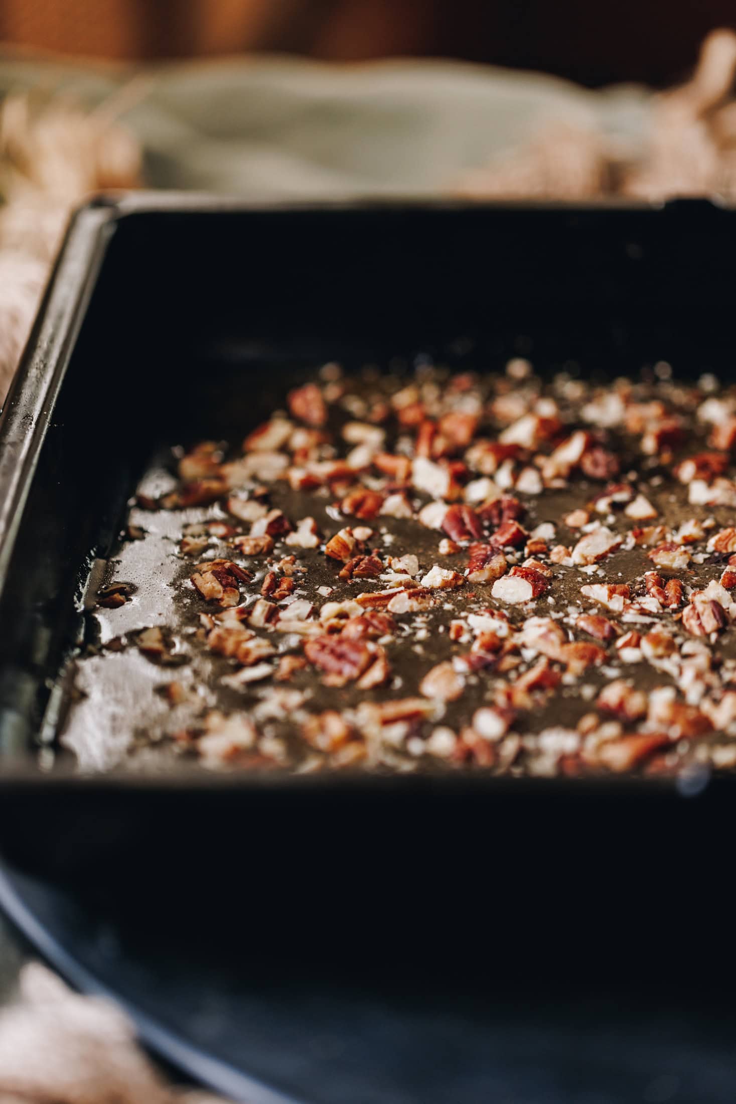 A black tray is sitting on a table limed with a green linen tablecloth. In the tray is gooey maple syrup caramel with chopped pecan nuts on top.