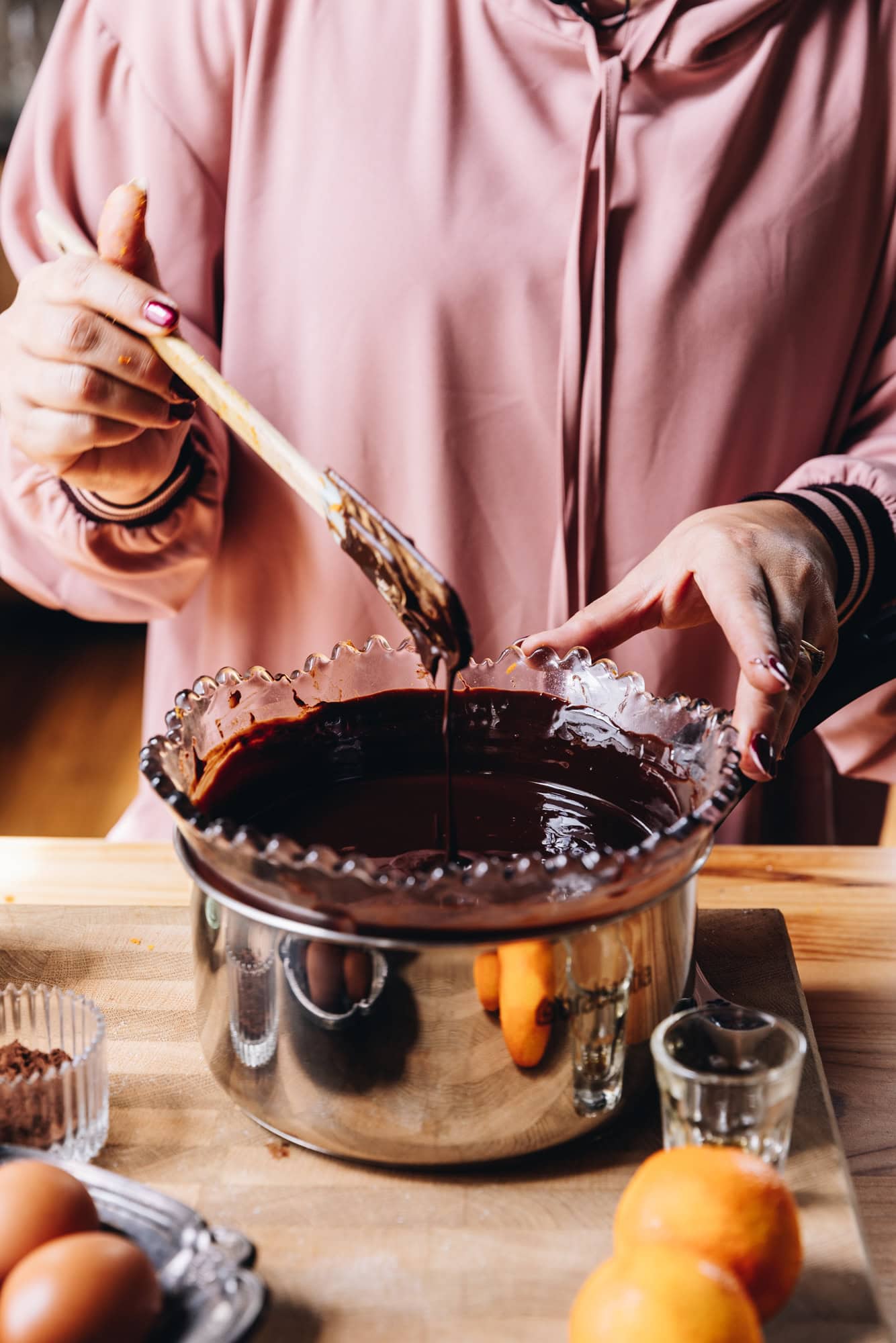 On a wooden table is a silver pot with a purple vintage bowl sitting inside it. Inside the bowl is melted chocolate. There are eggs, mandarins and vanilla around the pot on the wooden table. Naomi is holding the bowl and a wooden spoon behind it.