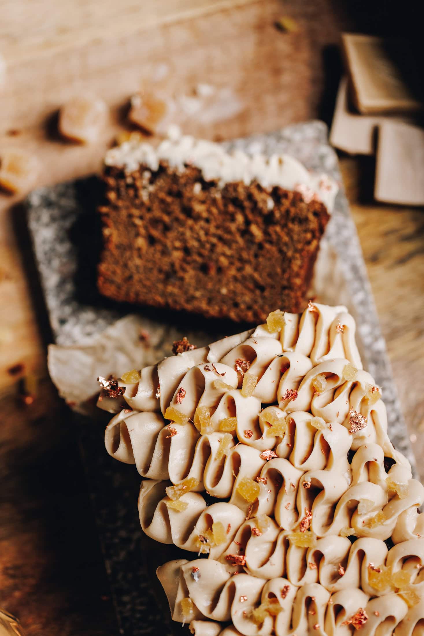 On a wooden table sits a small stone board. A top shot shows a baked spiced gingerbread loaf that has piped cream cheese frosting on it. It is sprinkled with crystallised ginger and speckles of gold leaf. 