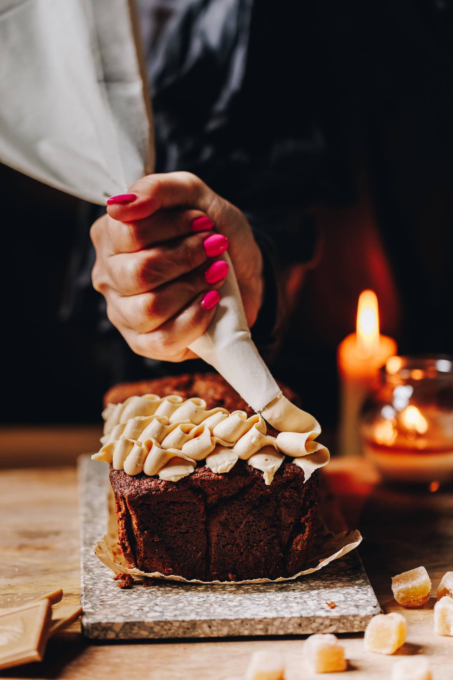 On a wooden table sits a small stone board. Two candles are burning in the background. On the stone board is a baked spiced gingerbread loaf that is having cream cheese frosting piped on it in a squiggly pattern. 