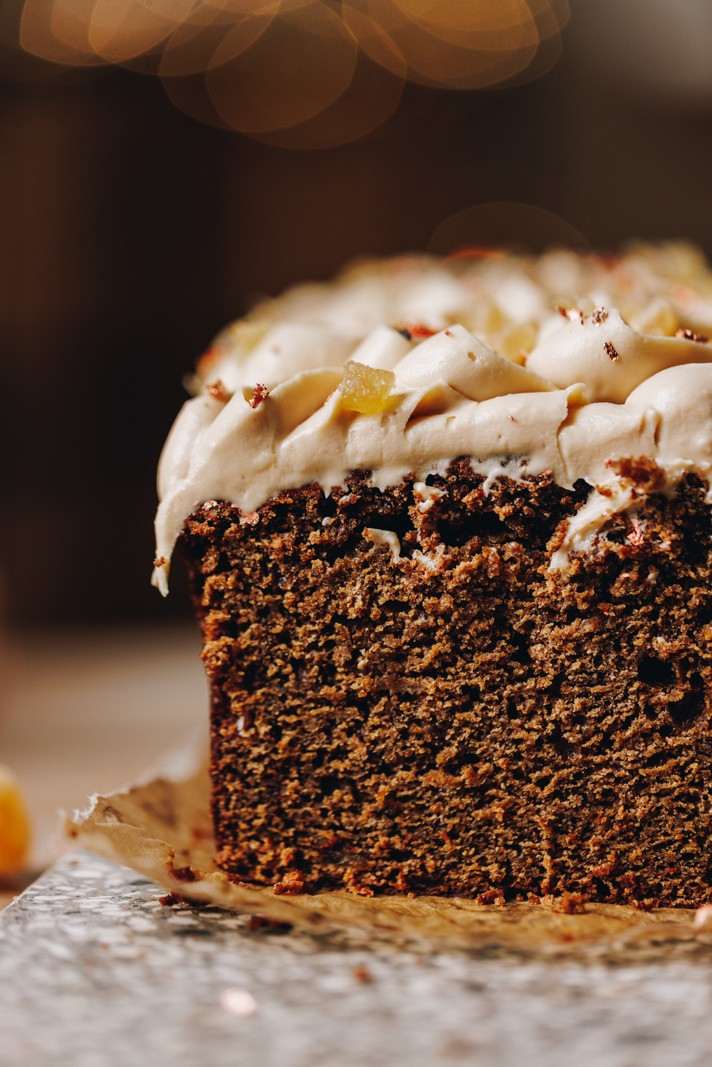 On a wooden table sits a small stone board. On the stone board is a baked spiced gingerbread loaf that has piped cream cheese frosting on it. It has been cut open to reveal the moist texture of the loaf. 