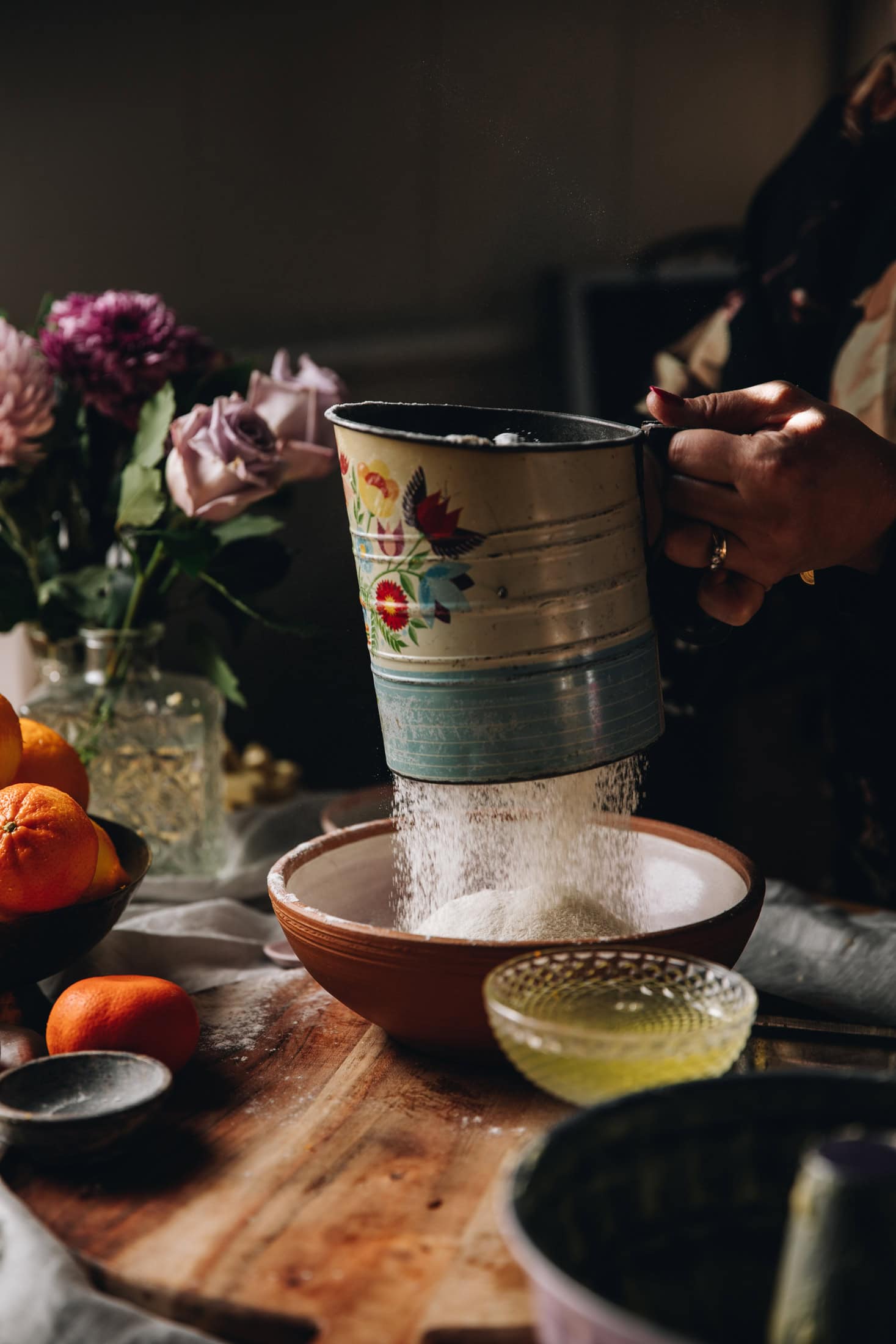 A wooden board has oranges sitting on it and a bunch of flowers in the background. A ceramic bowl sits on the wooden board and a vintage sifter is sifting flour in to it. 