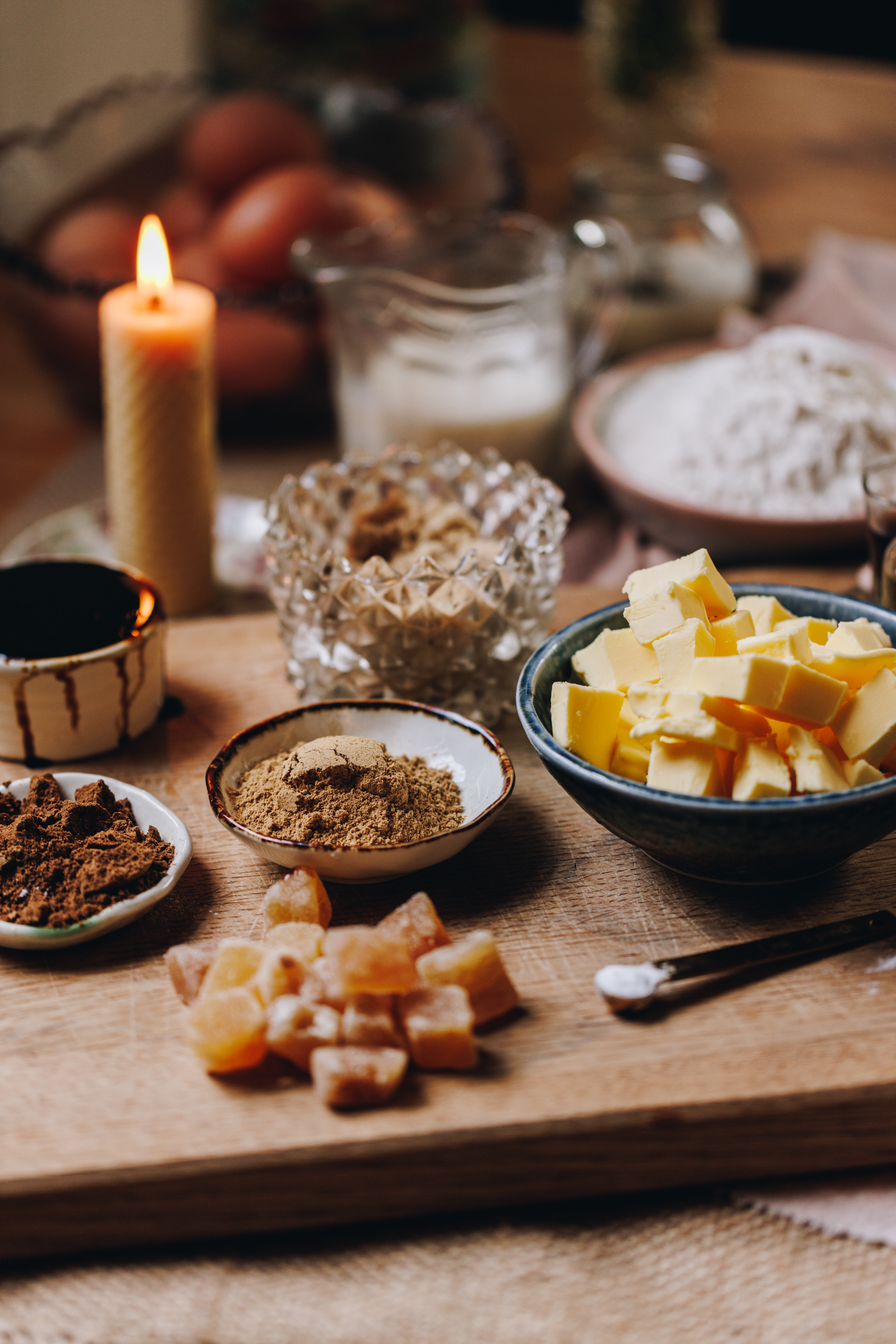 Cake ingredients are in different vintage bowls and are sitting on a wooden board on top of wooden table. There is a candle burning in the background. 