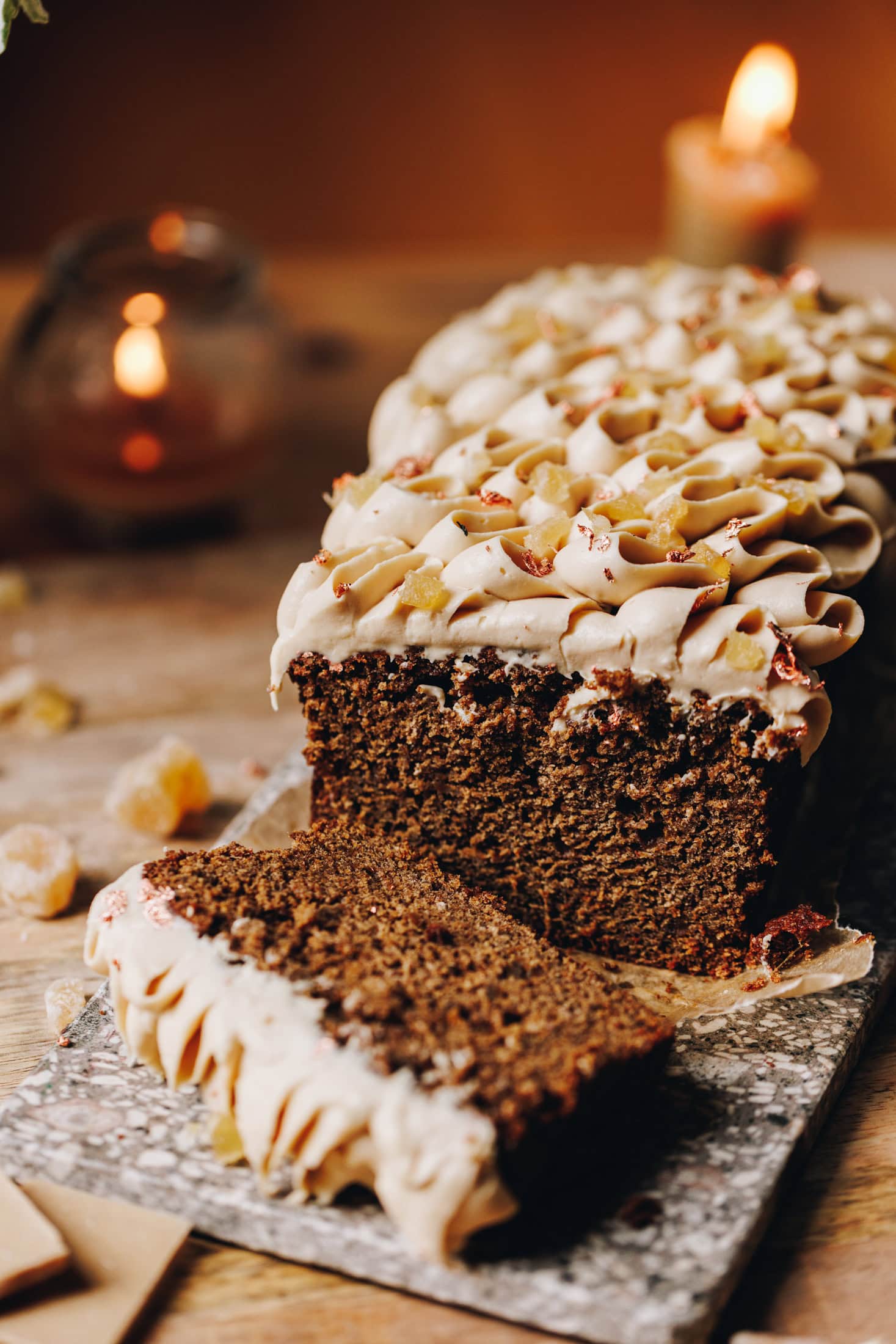 On a wooden table sits a small stone board. Two candles are burning in the background. On the stone board is a baked spiced gingerbread loaf that has piped cream cheese frosting on it. A lice has been cut and is laying flat, showing the inside of the loaf.