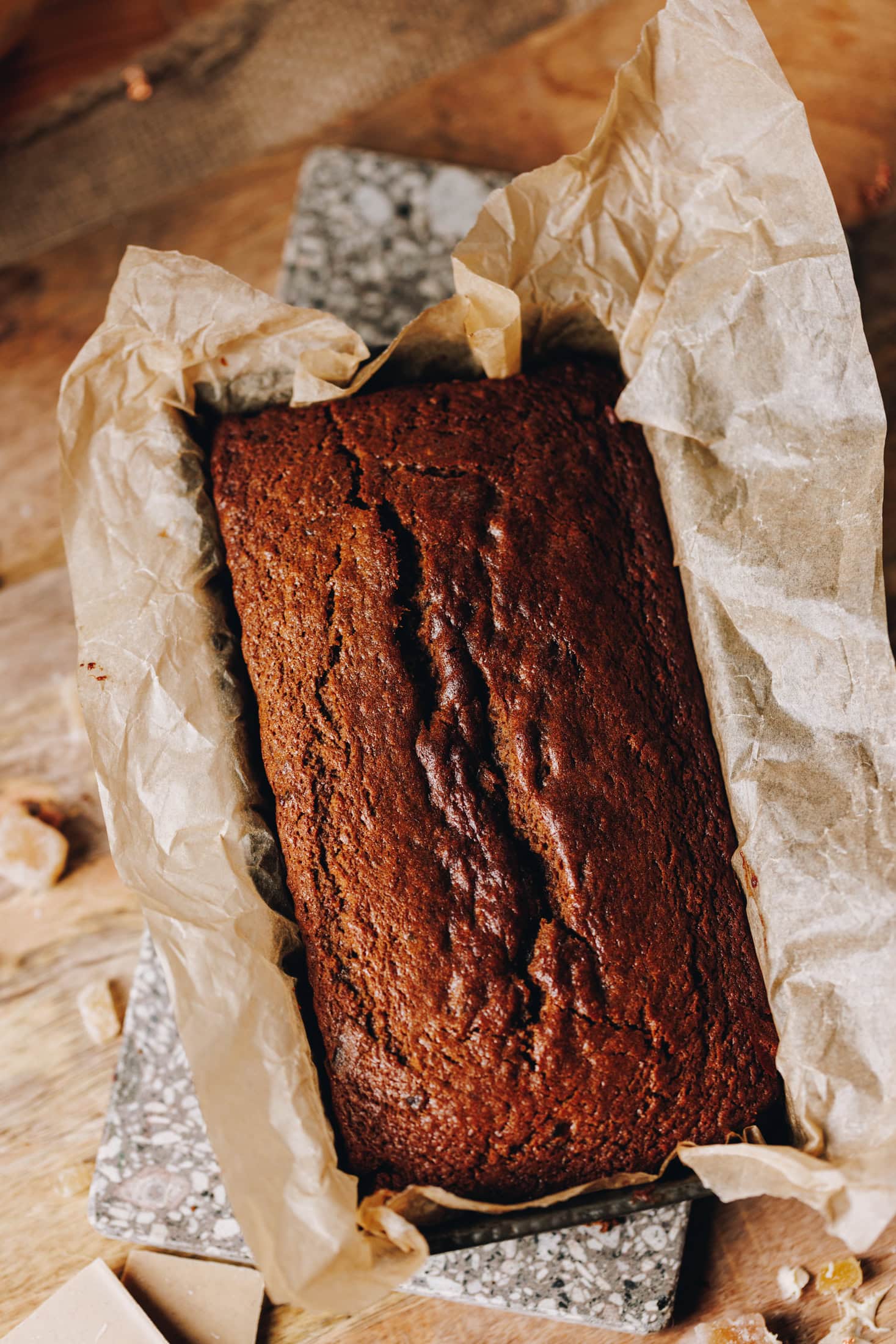 A top shot of a freshly baked spiced gingerbread loaf that is still in a tin lined with brown baking paper. It is sitting on a wooden table.