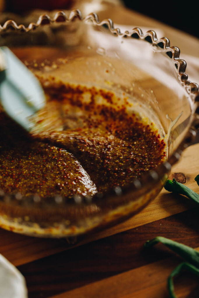 Honey and mustard is being stirred in a glass bowl with a blue spatula, it is an action shot. The bowl is sitting on a wooden board.