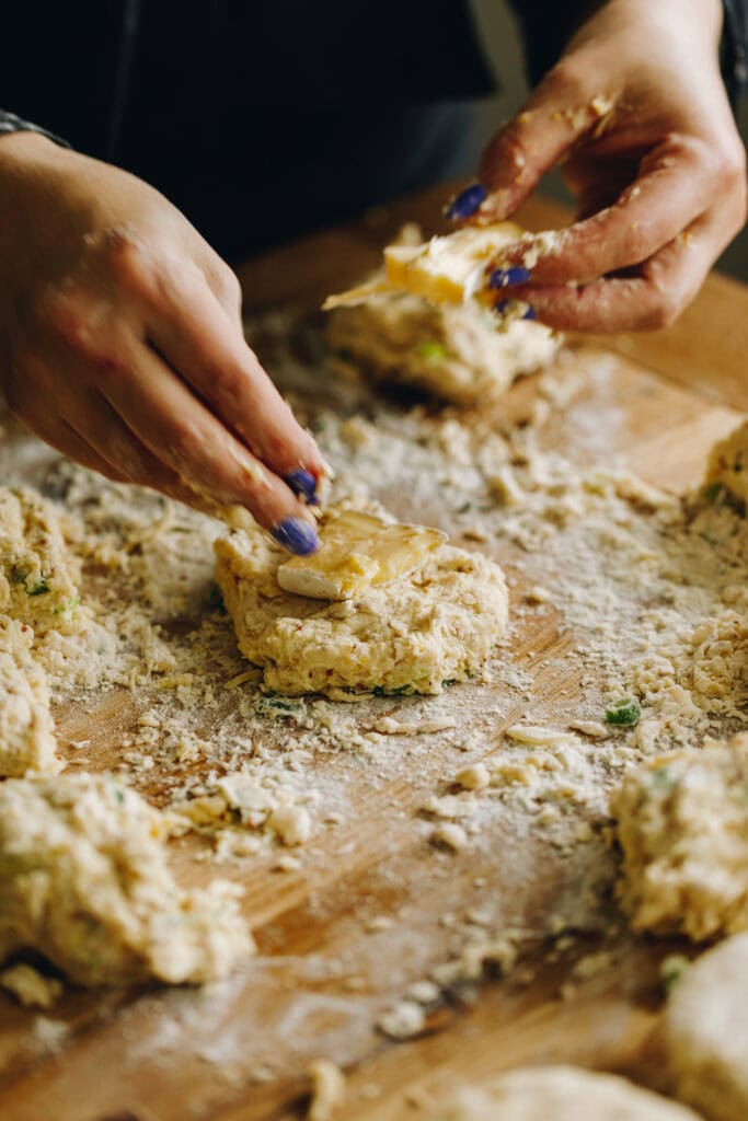 A piece of the Brie Stuffed Honey Mustard Scone dough is having brie placed on to it by a hand. It is sitting on a heavily floured, wooden bench.