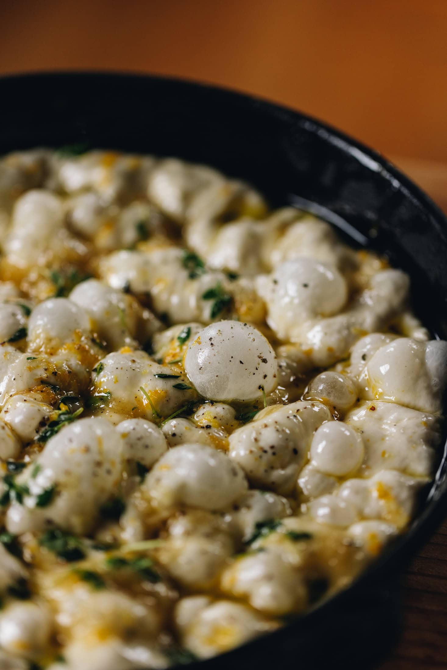 A close up of unbaked no-knead focaccia bread showing the bubbles that have formed in the dough. It is speckled with fresh thyme, lemon zest and garlic paste.