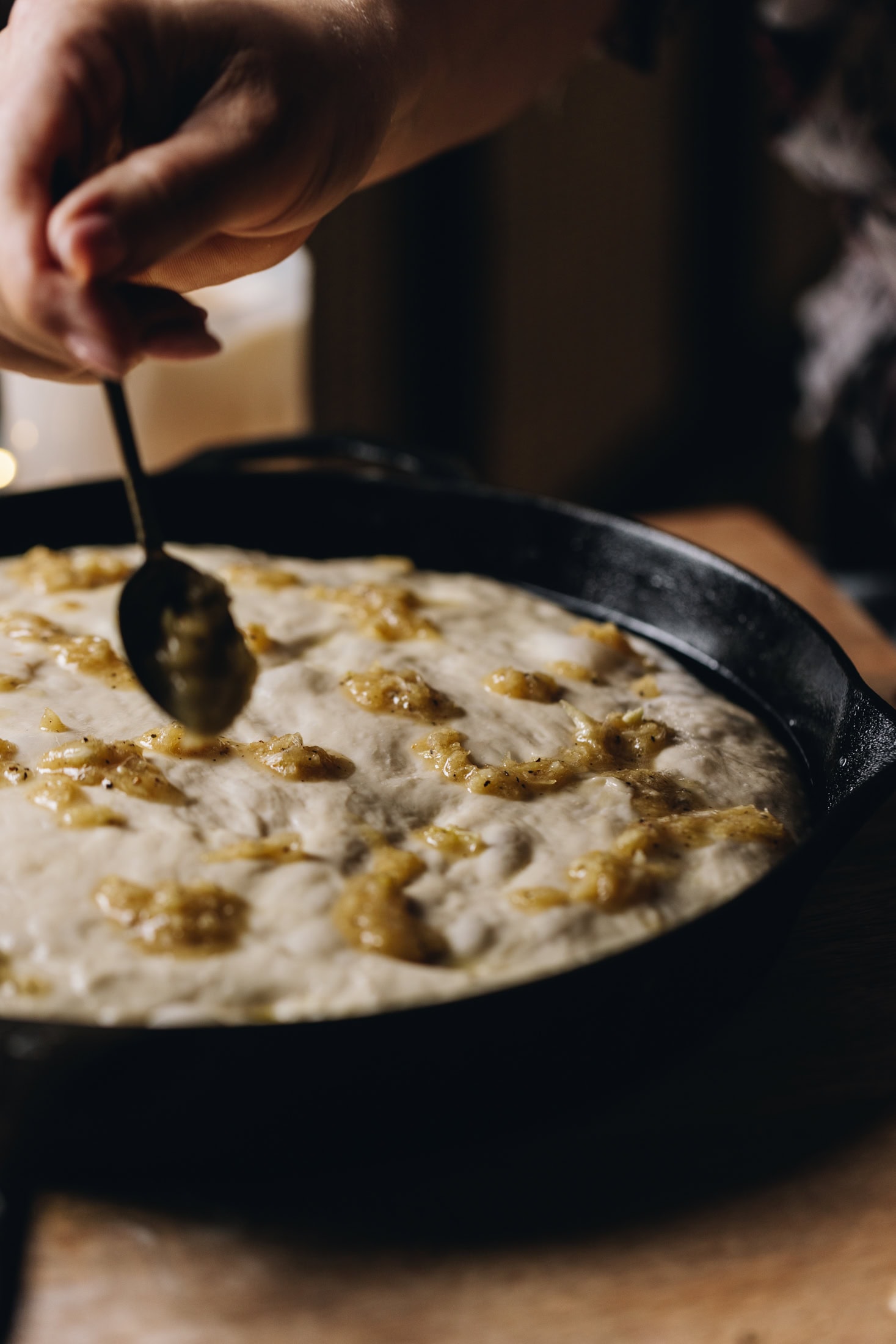 A black cast iron pan is sitting on a wooden table. In the pan is the no-knead focaccia bread dough that is unbaked. A teaspoon is adding garlic paste on top of it.