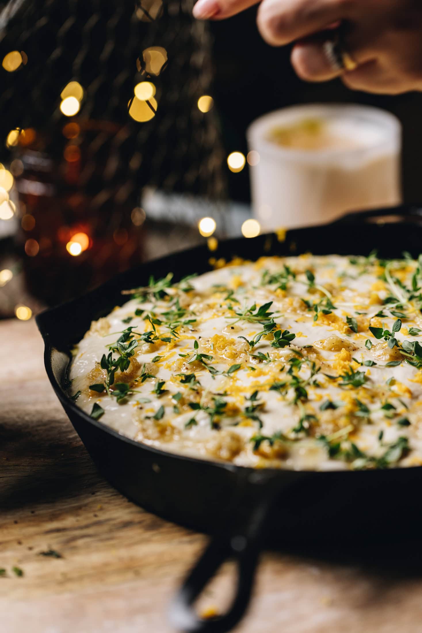 A black cast iron pan is sitting on a wooden table. In the pan is the no-knead focaccia bread dough that is unbaked. There is garlic paste, grated lemon zest and fresh thyme on top.