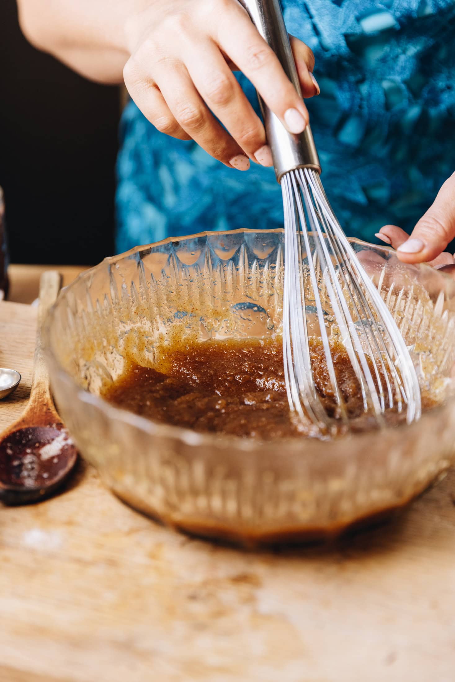 A glass bowl is sitting on a wooden table. In the bowl is brown butter and sugar being stirred with a whisk. 