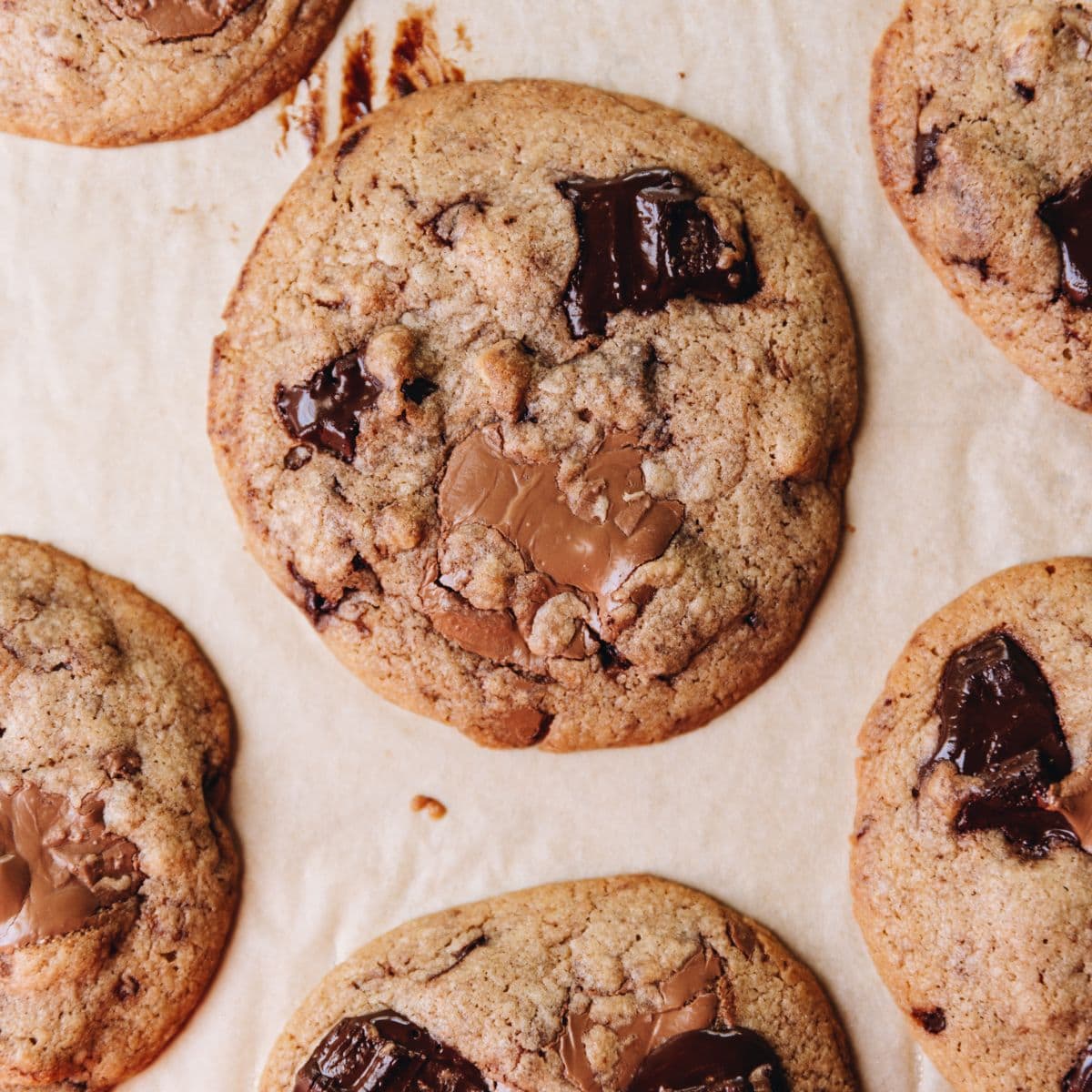 Freshly baked brown butter chocolate cookies are on brown baking paper. The flay lay shot shows a close up of a cookie with melty dark and milk chocolate chunks on it.