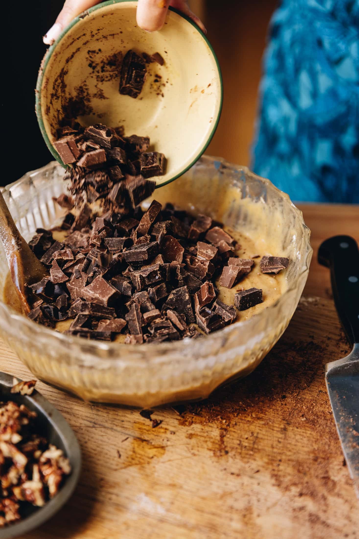 Chopped milk and dark chocolate is being added in to the gooey cookie dough mixture. The chocolate is dropping in from a small vintage bowl. The bowl is on a wooden table. 