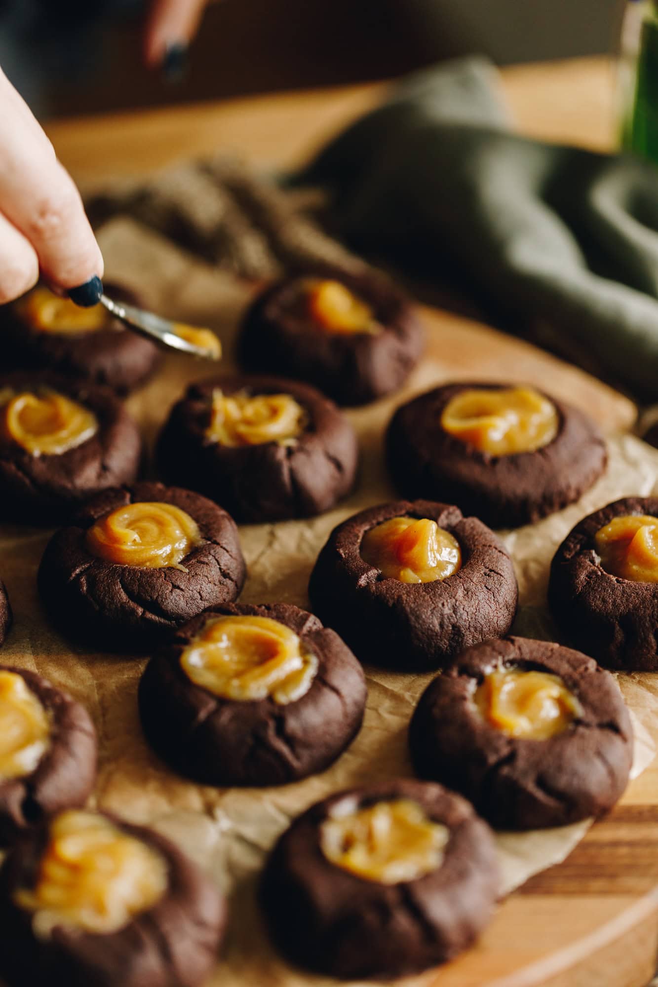 On a wooden table is a tray lined with baking paper that is filled with chocolate cookies that has thickened easy caramel sauce in each one. 