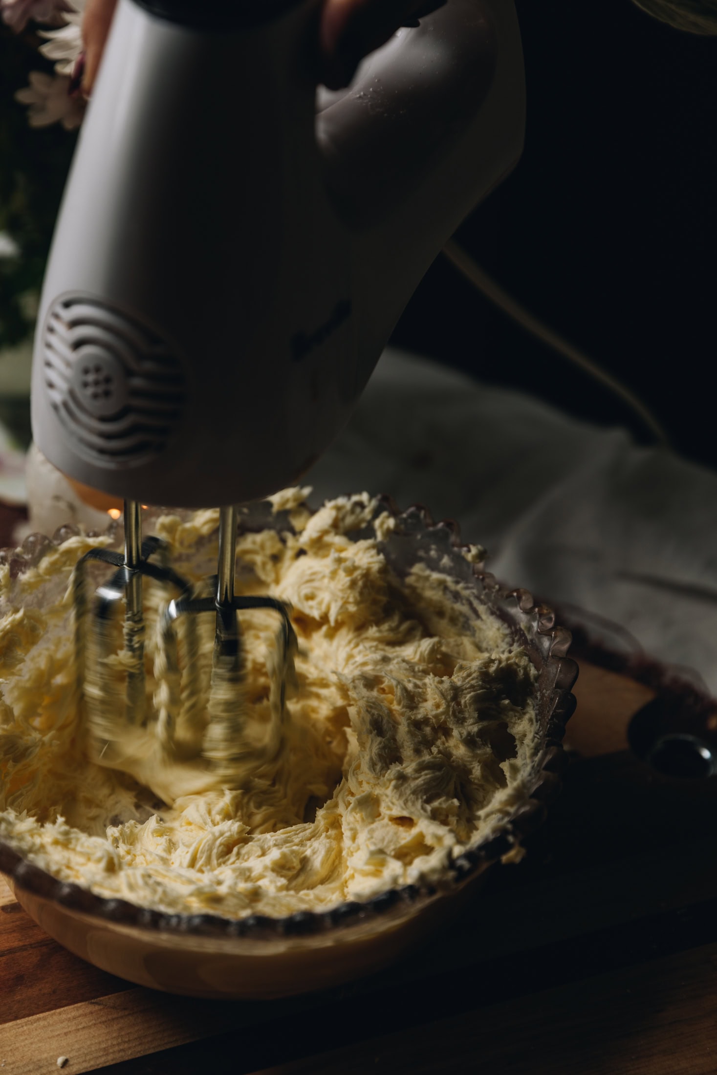 In a vintage glass bowl is cream cheese frosting being whipped with a  white hand mixer. It is sitting on a wooden board.