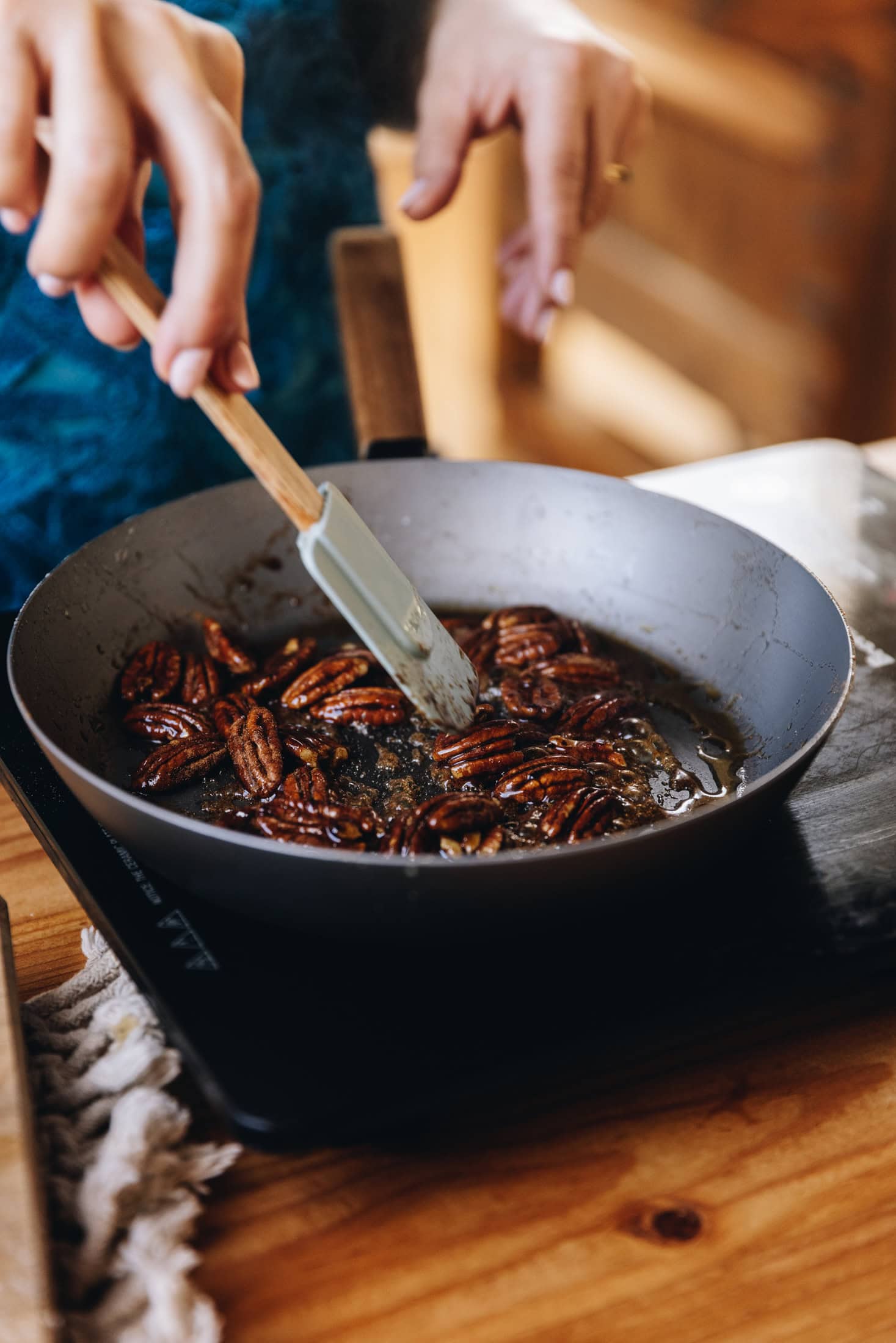 Spiced pecan nuts are being stirred with a spatula in a fry pan, it is sitting on a portable stove on a wooden table. 
