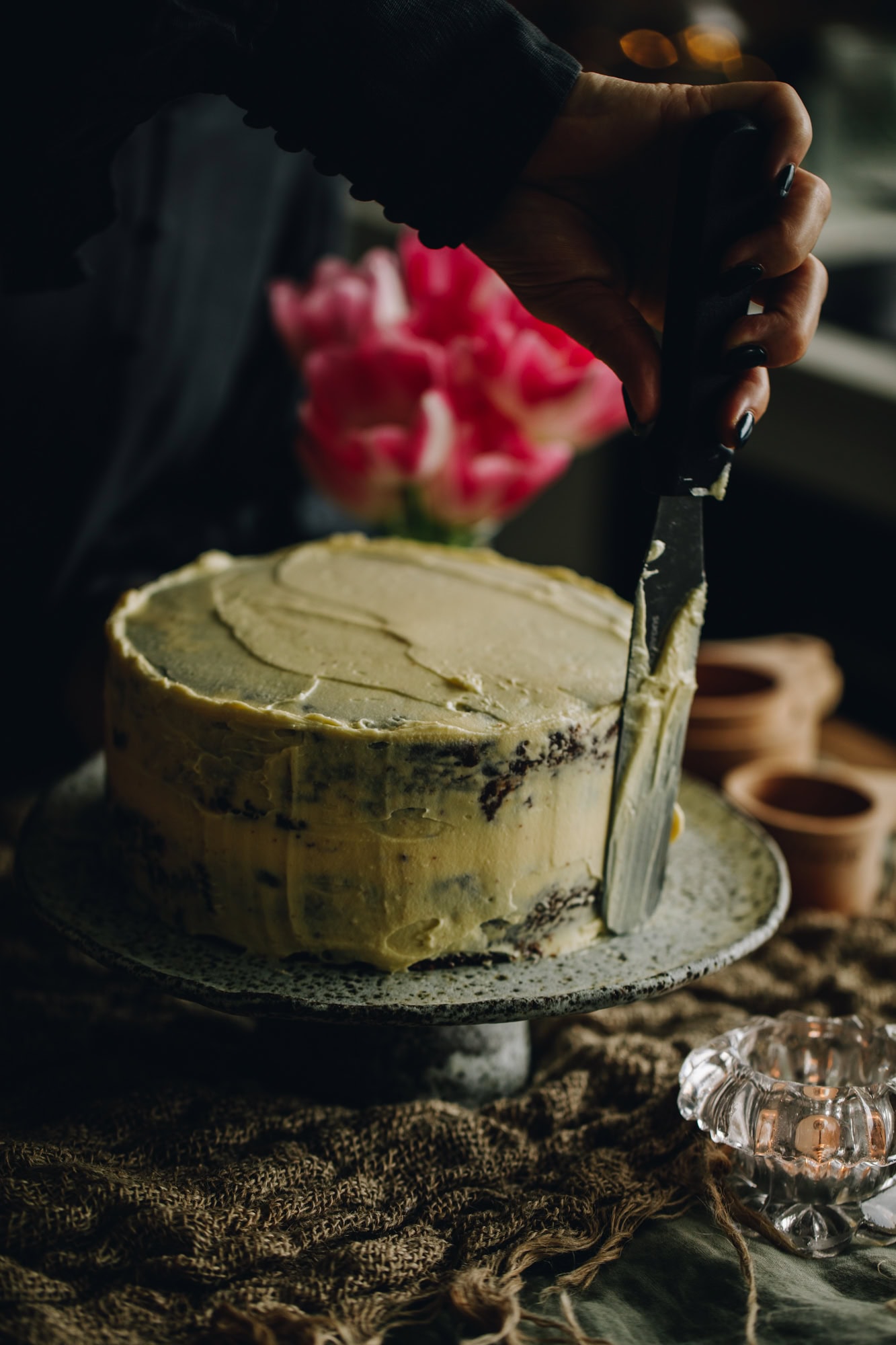 A ceramic cake stand sits on a wooden table with a bunch of flowers in the background. On the cake stand is a Sticky Caramel Date Cake with cream cheese frosting being spread on to it with a offset spatula.