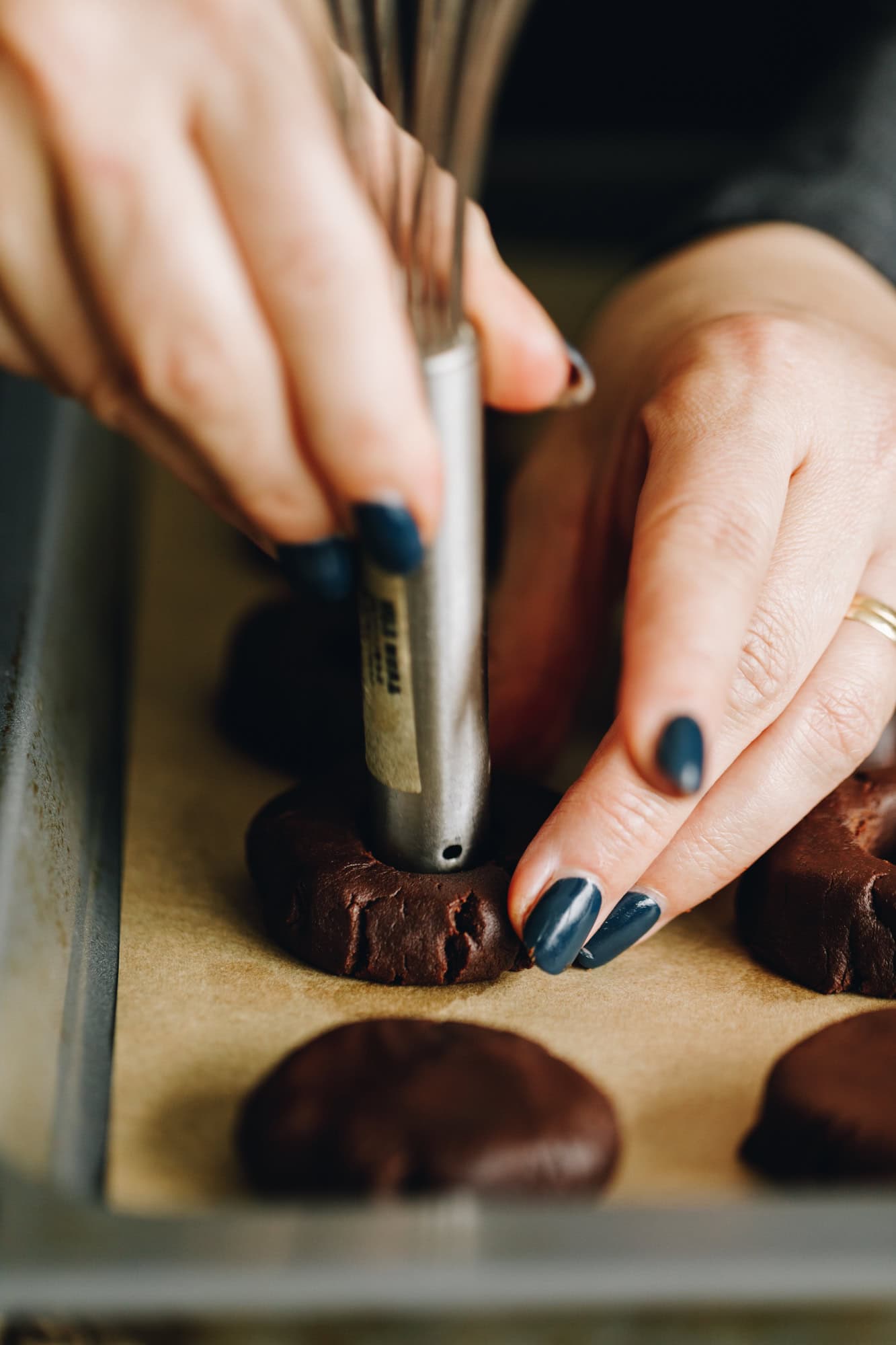 On a tray with brown baking paper are unbaked chocolate cookies that have been flattened down. Naomi is indenting one with a silver handle. 