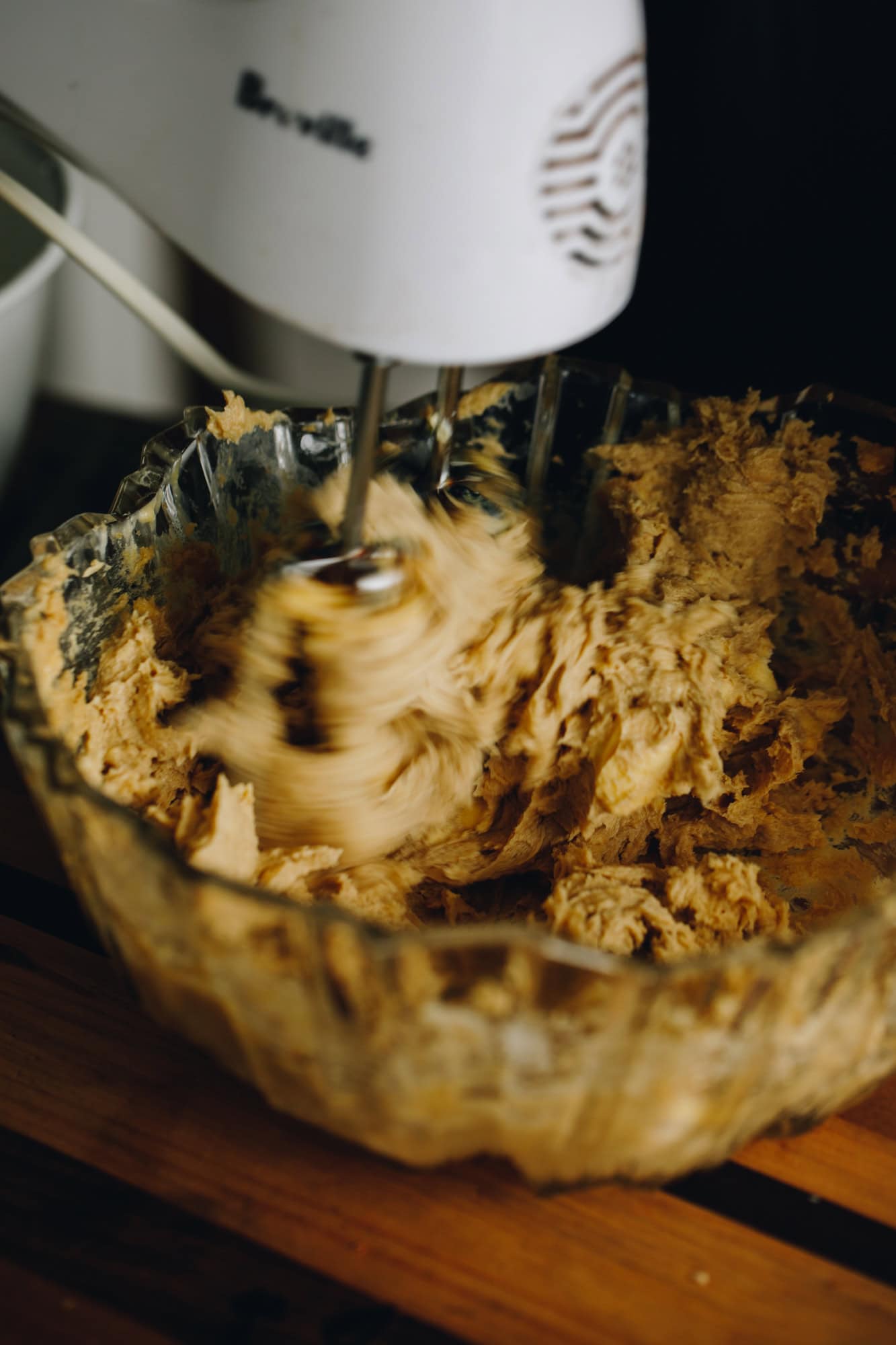 In a vintage glass bowl is butter and sugar being whipped with a white hand mixer. It is sitting on a wooden board.