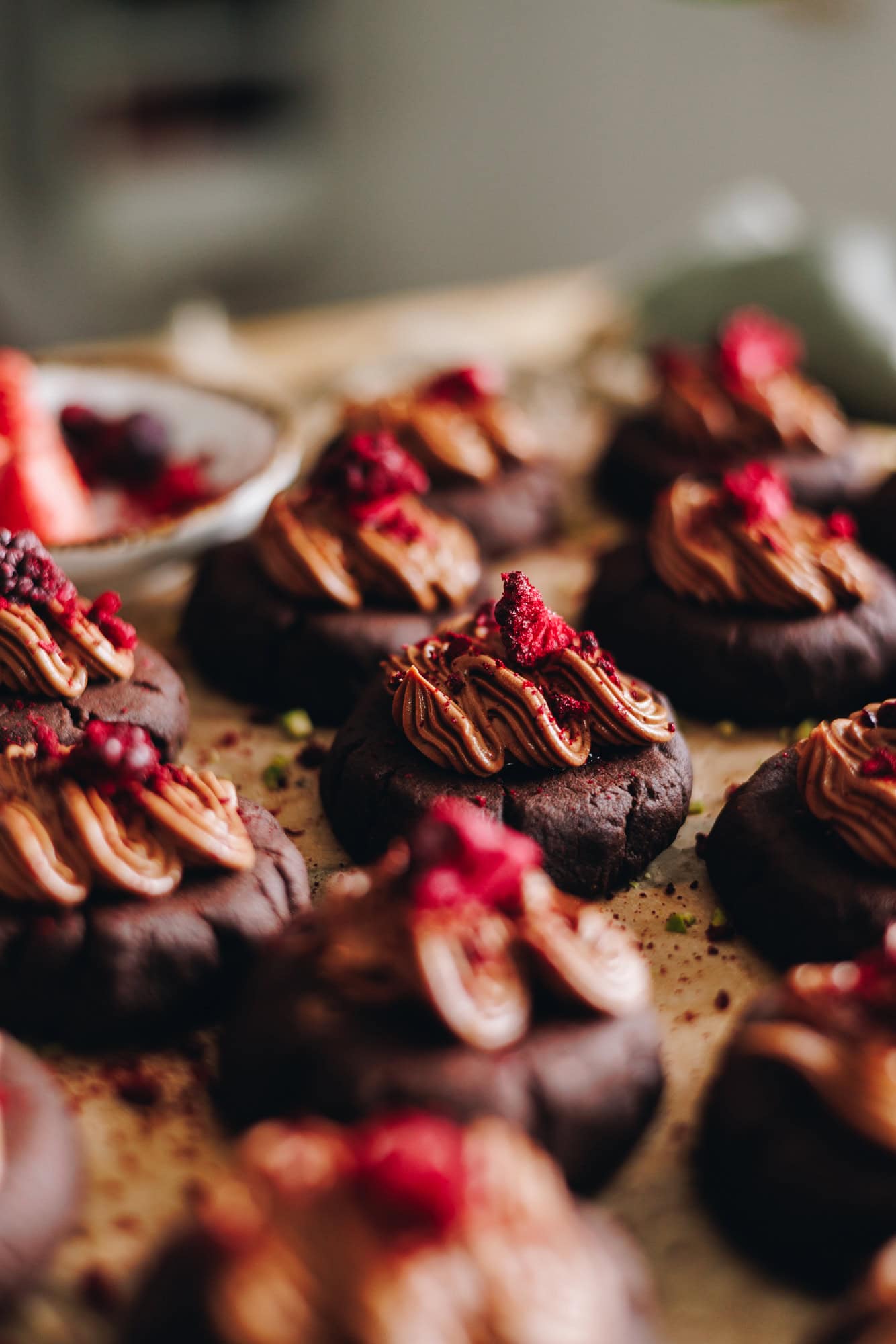 On a wooden board that is covered in brown baking paper sits on a table. On top are decorated chocolate and raspberry cookies. They have chocolate frosting on top of each one and freeze-dried boysenberries sit on top.