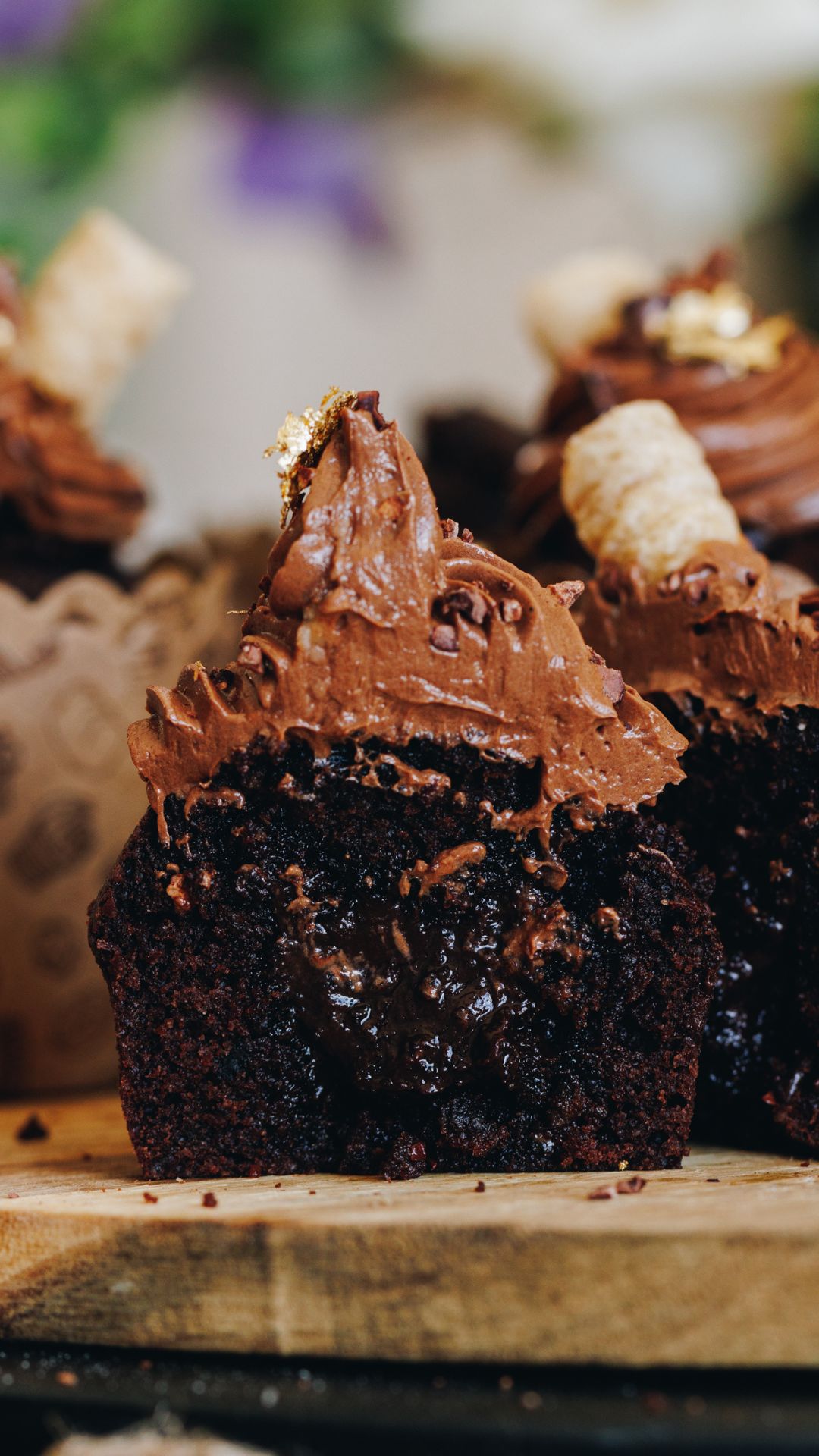 Chocolate ganache  muffins sit on a wooden board. One of the muffins is in focus and it has been cut in half to reveal the gooey chocolate centre and the chocolate frosting piled on top. 