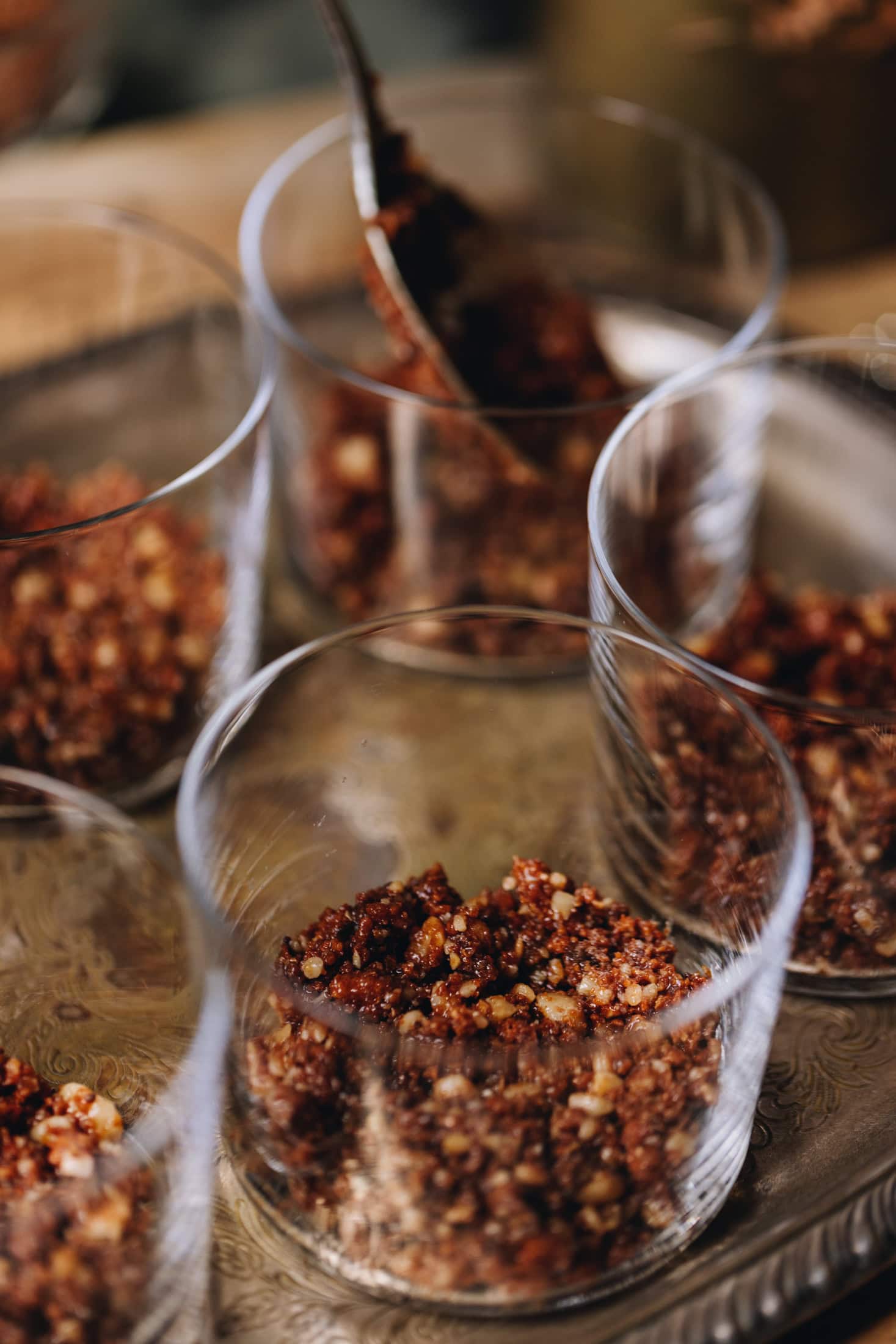 Small glasses are on a golden tray, in each of them are biscuit crumbs, a spoon can be seen pressing it down. 