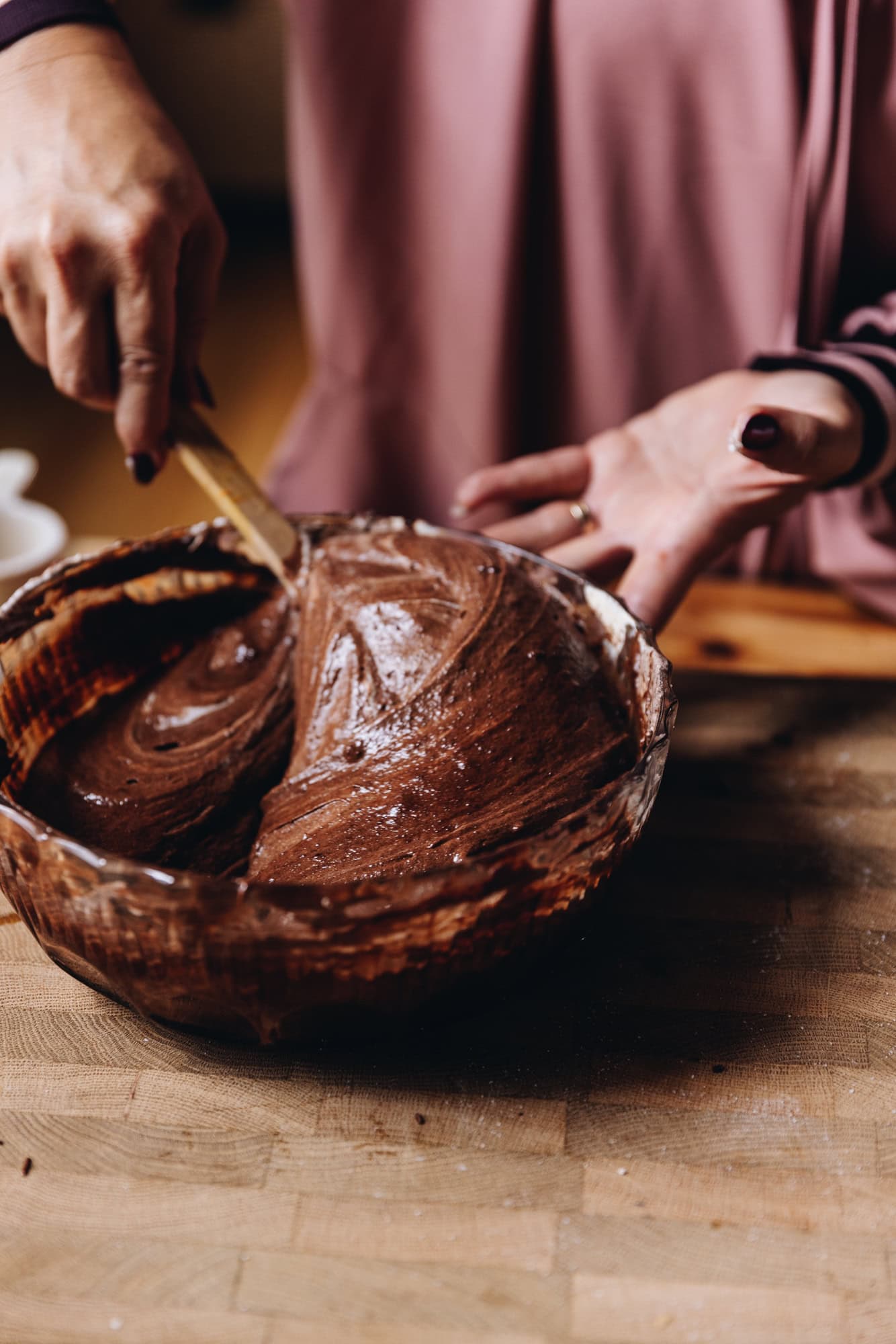 On a wooden table is glass vintage bowl. Inside it hands are folding a thick chocolate mixture. 