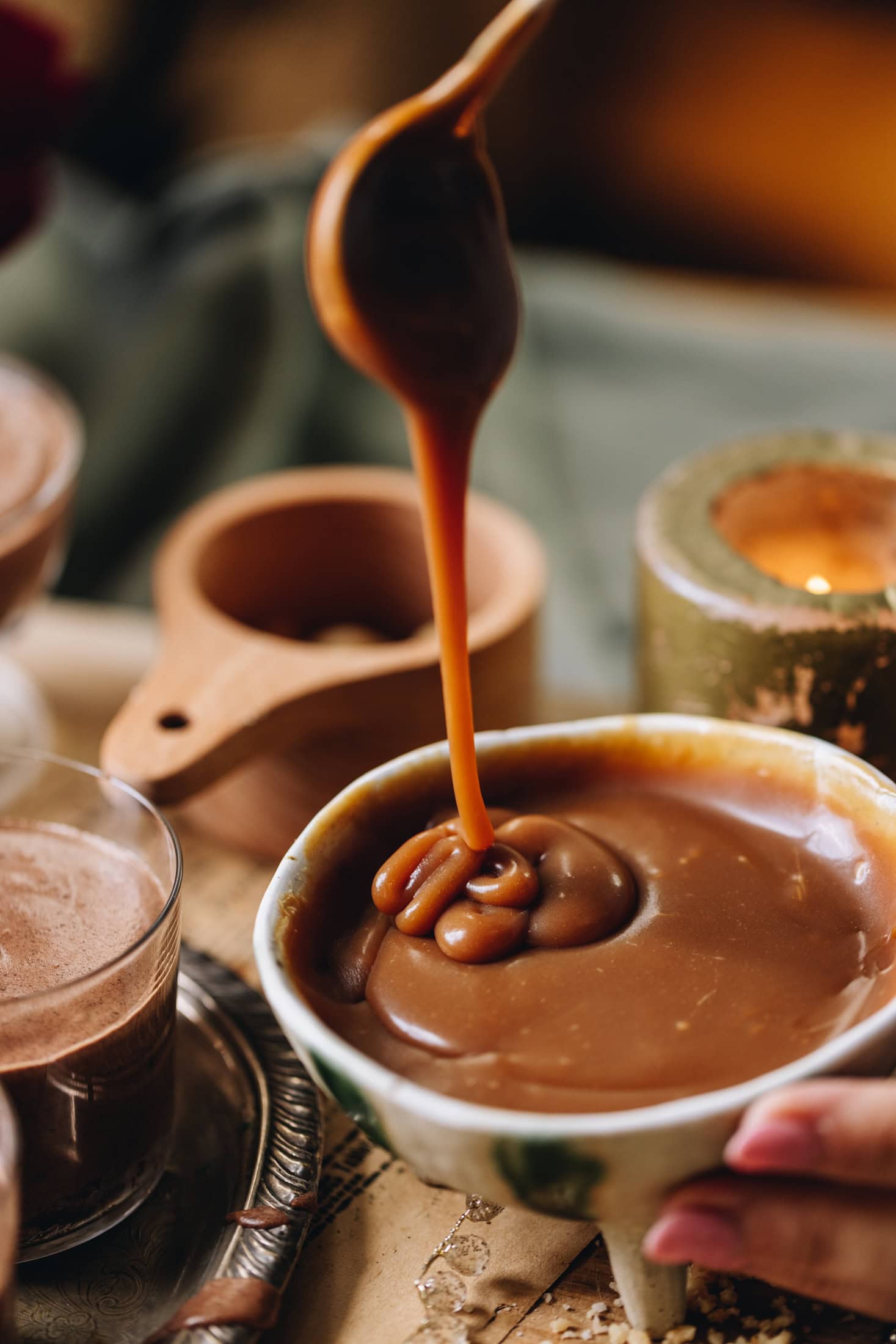 A hand is holding a ceramic bowl with thick salted caramel in it, a spoon is drizzling the sauce back in to the bowl revealing the smooth texture. 