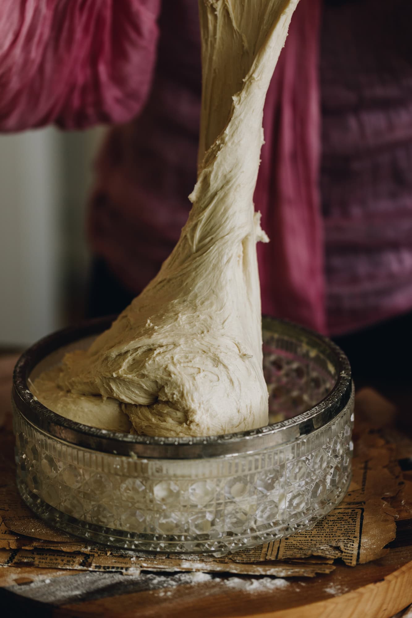 No-knead bread bun dough is being stretched and folded. The dough is being lifted high out of the vintage glass bowl. It is on a wooden board. 