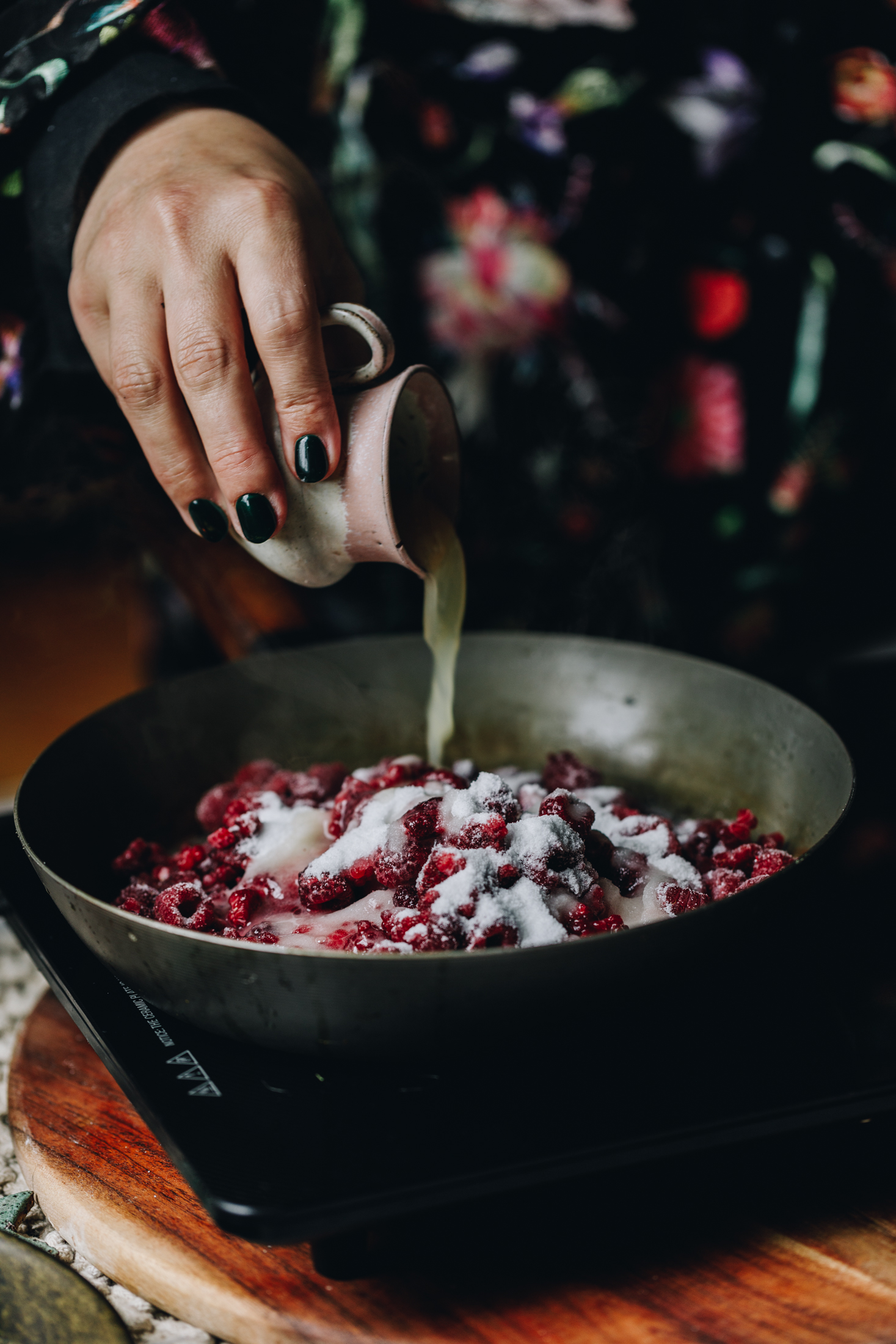 A moody shot shows a frying pan on top of a portable stove. It is on a wooden table. In the pan is frozen raspberries and white sugar. A small ceramic, pink jug pours in the lemon juice in to the centre of the pan.