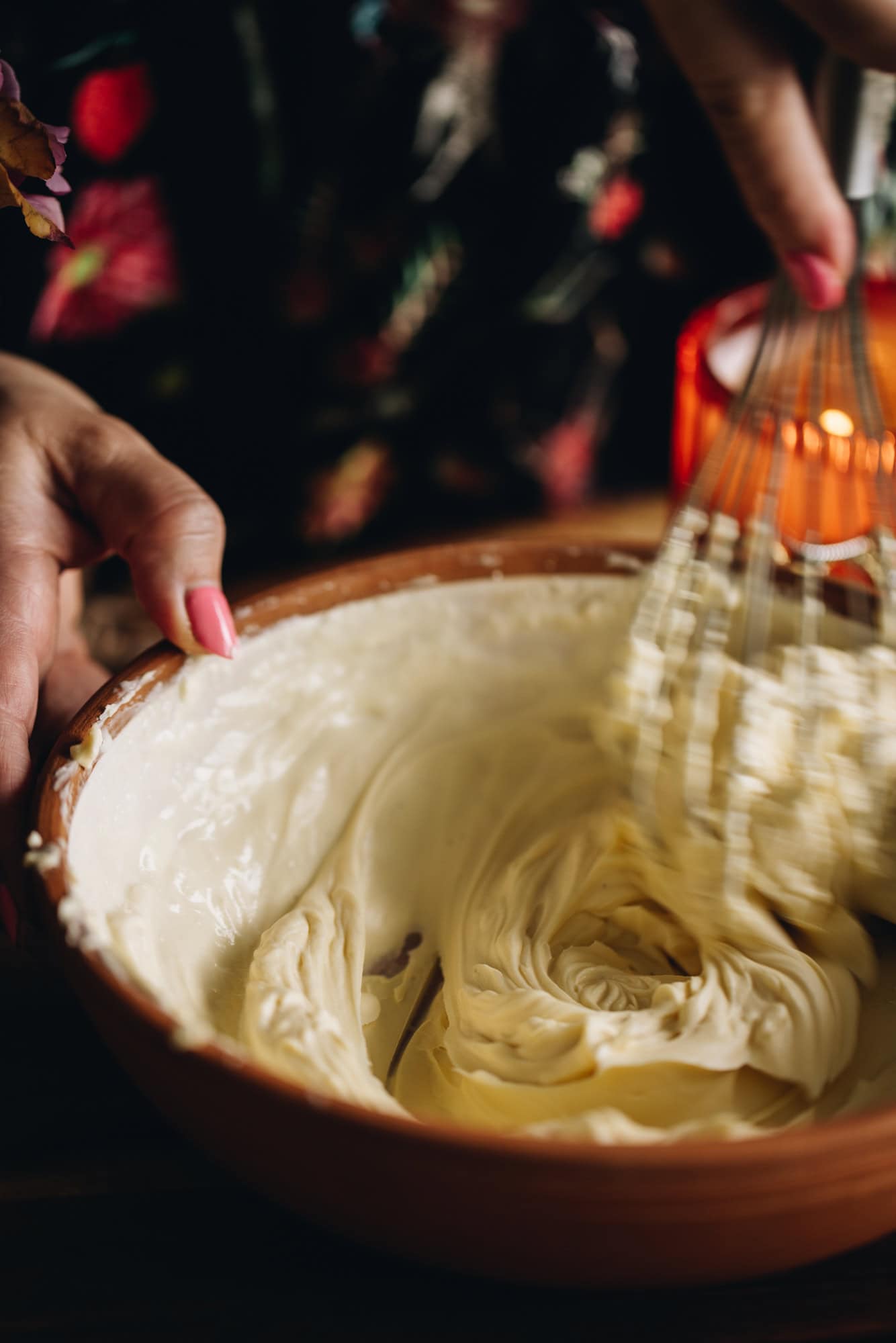 In a ceramic bowl is cream cheese frosting that is being whipped with a wire whisk. There is an orange candle in the background. 