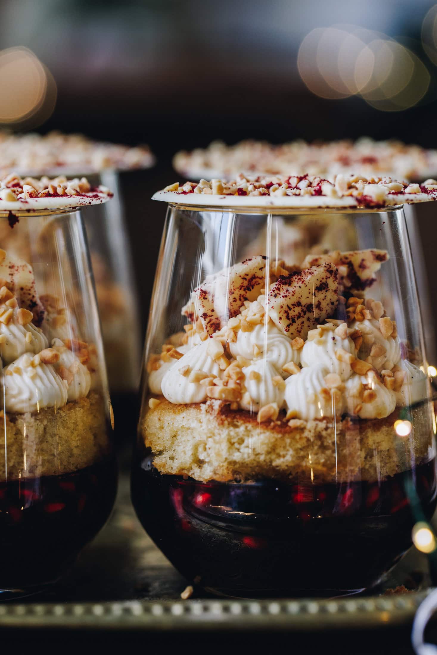 A close up shot shows the Mini Berry Trifles that has been decorated and set in glasses. It shows a pomegranate jelly, a sponge, piped whipped cream, nut, white chocolate shards. The glass is topped with a white Chocolate disk.